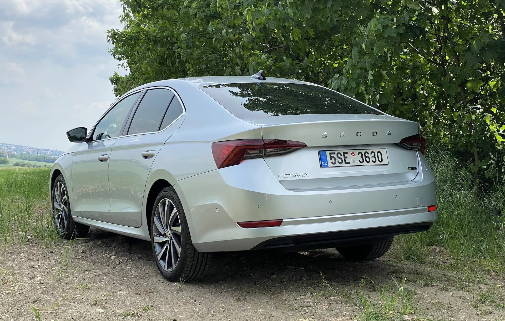 Hátulról háromnegyedes nézetből látható egy világos színű 2020-as Skoda Octavia szedán földes felületen, háttérben fűvel és fákkal. A fotó kiemeli a jármű hátsó és oldalsó profilját, sima vonalakkal, kontúros felületekkel, könnyűfém felnikkel és elegáns LED hátsó lámpákkal. A Skoda embléma és az 'OCTAVIA' modellnév jól látható a csomagtérfedélen, valamint egy európai rendszámtábla. A természetes, lágy fény kiemeli az autó tervezési elemeit, anélkül, hogy éles árnyékokat keltene, ami borús égboltot vagy szűrt napfényt sugall.