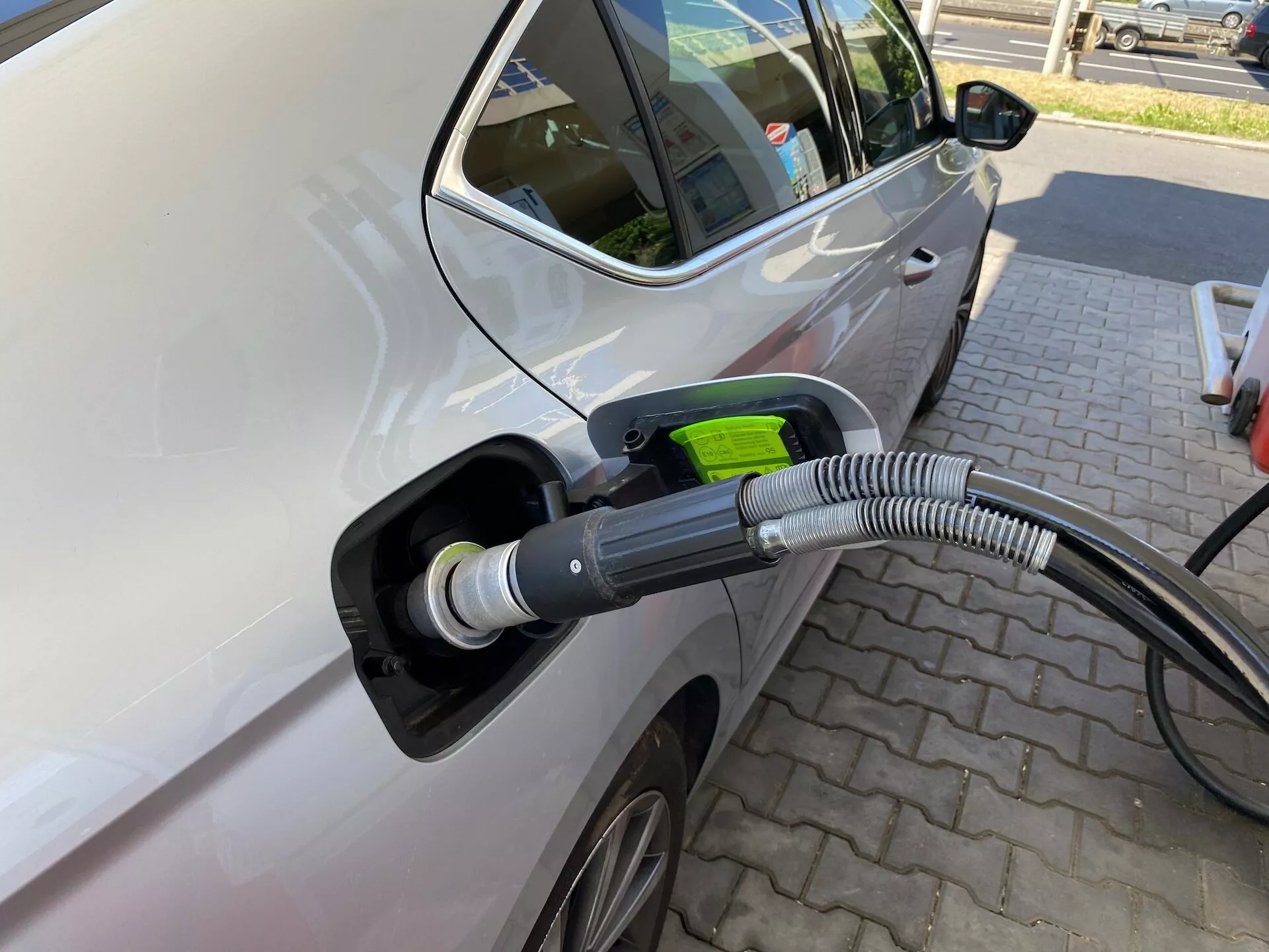 A Skoda Octavia 2020 bal oldalának közeli képe, amely egy elektromos töltőállomáshoz csatlakozik, kiemelve a töltőport területét, a töltőkábelen és a porton fókuszálva. A kép enyhe szögből van készítve az elejétől a hátul felé, bemutatva az első vezető oldali ajtót és a hátsó ajtó egy részét, jelezve, hogy több ajtós járműről van szó. A jellemzők közé tartozik a modern oldalsó tükör, részben látható alufelni, világos színű autó fényes felülettel, nyitott töltőport vastag, hullámos védőburkolattal a kábelen, valamint zöld színű csatlakozófogantyú, amely az ökobarát természetét szimbolizálja. Az autó egy mintázatos téglás burkolatú területen parkol, ami egy dedikált töltőállomást sugall, más járművek nélkül a közelben, ezzel hangsúlyozva a töltési műveletet.