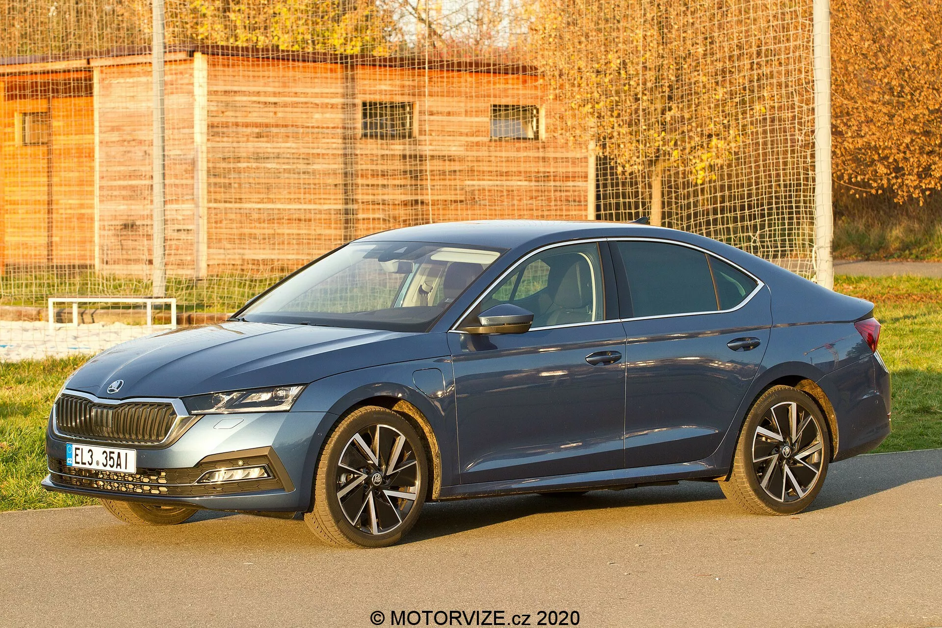 A 2020-as Skoda Octavia négyajtós szedán képének háromnegyed elülső nézete, amely bemutatja az elülső rácsot, a fényszórókat, a motorháztetőt, a kerekeket, a vezető oldali ajtót és a hátsó rész egy részét. A felvétel megörökíti a rács jellegzetes kialakítását, a valószínűleg LED vagy adaptív világítórendszerekkel felszerelt elegáns fényszórókat, a sportosságot vagy luxust sugalló, többrészes alufelnikkel ellátott kerekeket, az alacsony profilú gumiabroncsokat és az irányjelzőkkel integrált oldalsó tükröket. A jármű aerodinamikus vonalai és kék színe jól látható, kiemelve a design és technológiai jellemzőket, amelyek az autóipar és modern járművek kontextusában megfelelőek.