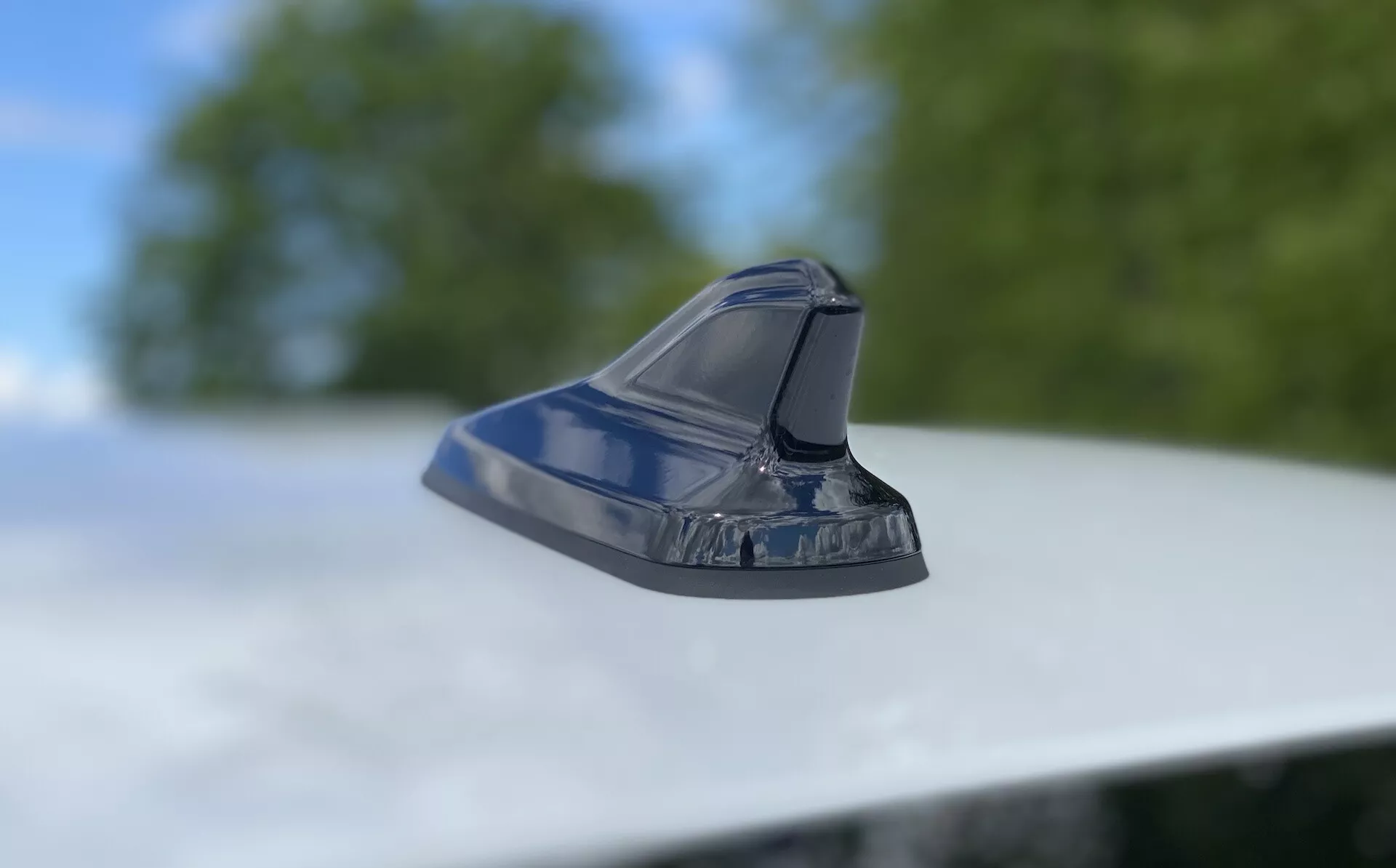 Közeli kép egy cápauszony stílusú antennáról egy jármű tetején, bemutatva a modern autóipari dizájnt. Alacsony szögből készült felvétel, a kamera enyhén felfelé döntve, az antennát fókuszálva, amely egy aerodinamikus, fényes formát mutat elmosódott lomb- és égbolt háttér előtt. Az áramvonalas kialakítás FM/AM rádió-, műholdas navigációs- és fejlett kommunikációs technológiákra utal. A fényes bevonat tükrözi a jármű jól karbantartott külsejét.