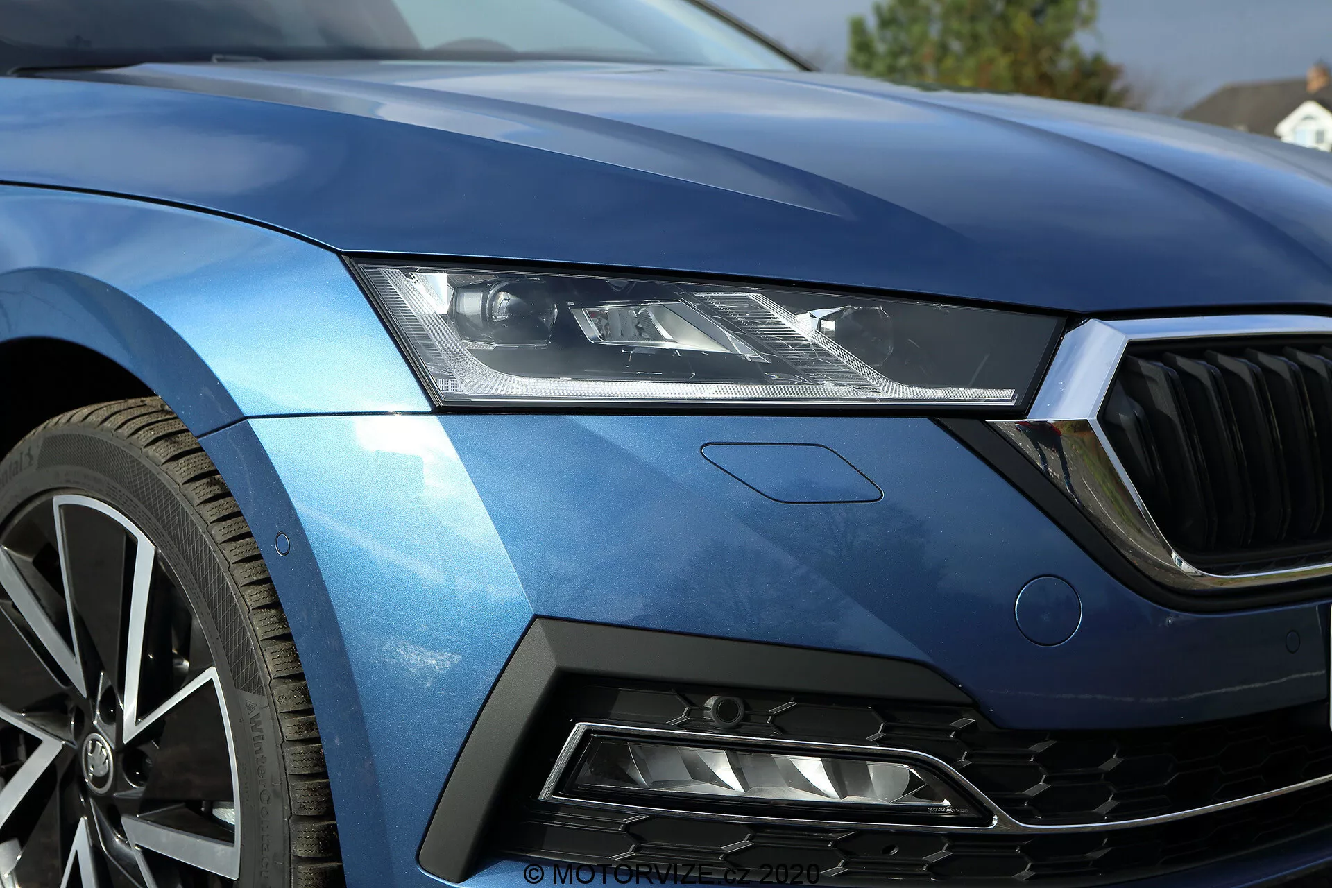 Közelkép egy 2020-as Skoda Octavia limuzin első bal negyedéről, amely az elegáns LED fényszórókat, a motorháztető egy részét, az első hűtőrács bal szélét, az aerodinamikus bal első sárvédőt és a lökhárító egy szegmensét, amely méhsejtrácsú légbeömlővel van ellátva, mutatja. A fémes kék festés és az éles vonalak a luxust és a sportosságot hangsúlyozzák. A többrészes könnyűfém kerék magas teljesítményt sejtet. A design egy finom jelvényt is tartalmaz, amely egy adott felszereltségi szintet vagy technológiai csomagot jelez. Ez a kép gyönyörűen megörökíti a jármű fejlett világítástechnológiáját, a festés felületét és a teljesítményorientált tervezési jellemzőket.