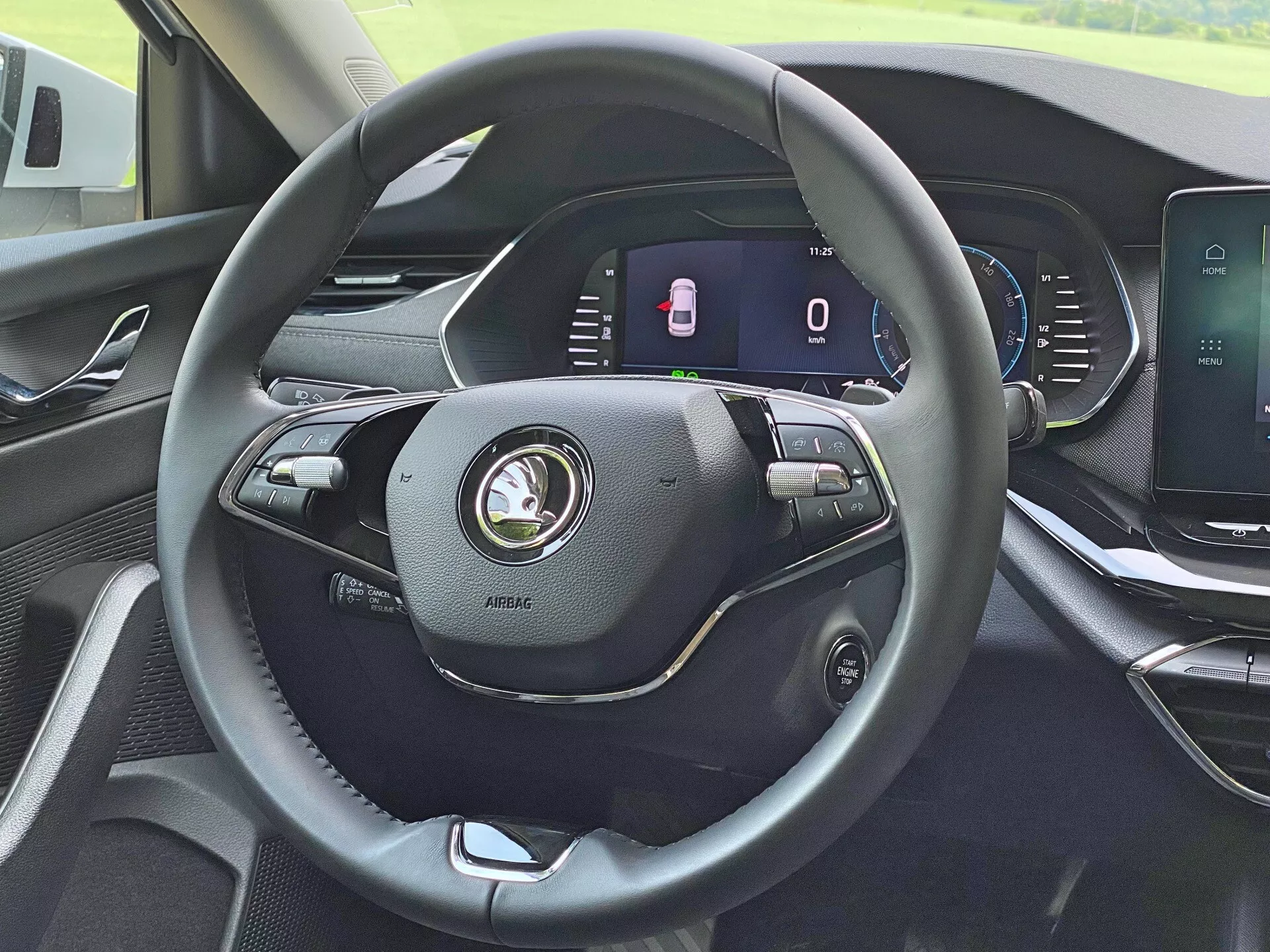 A Skoda Octavia Limousine 2020 belső tere vezetői szempontból, kiemelve a modern, háromküllős bőrborítású kormánykereket, amely különféle vezérlőkkel rendelkezik hangerőszabályzáshoz, hangvezérléshez és sebességtartó automatikához. A kormánykerék mögött digitális műszerfal látható, amely álló sebességmérőt és nyitott ajtók jelzőjét mutatja. A középkonzolon egy érintőképernyős infotainment kijelző található multimédiás funkciókhoz, navigációhoz és járműbeállításokhoz. A műszerfal modern formatervezésű és kiváló minőségű anyagokkal készült.