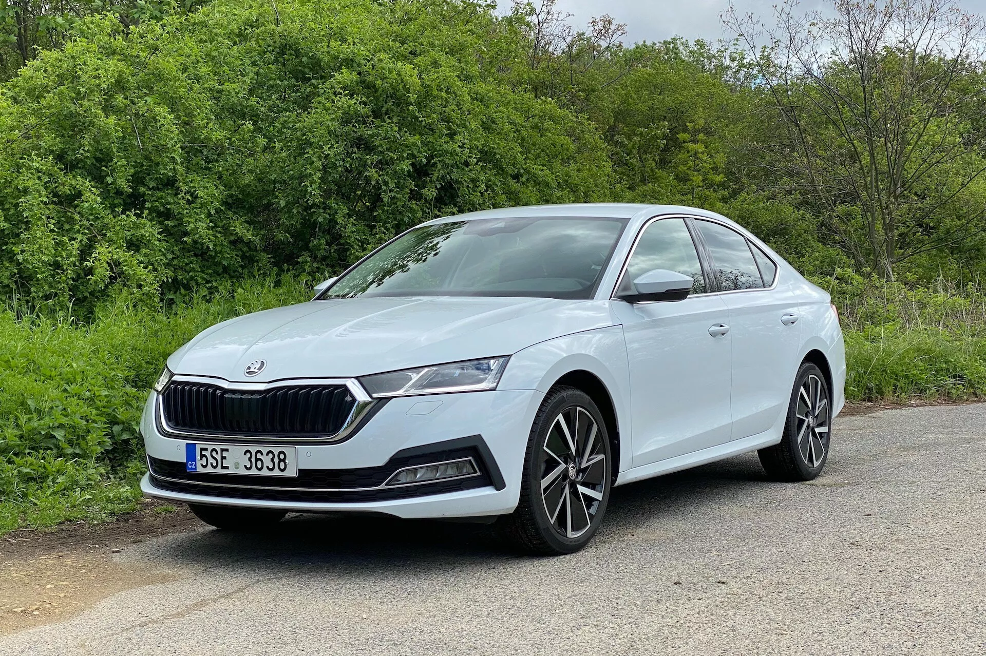 A kép egy 2020-as Skoda Octavia szedánt ábrázol, háromnegyed elülső szögből nézve, megjelenítve az első homlokzatot és a vezető oldal-profilját. Az autó aszfaltos felületen parkol, háttérben természetes elemekkel, mint fák vagy bokrok, jellegzetes rácsot, elegáns LED fényszórókat, ködlámpák fészkeit és légbeömlőket mutatva a lökhárítón. A többrudas könnyűfém kerekek és az oldalsó, gördülékeny, aerodinamikus vonalak fokozzák a modern és sportos megjelenést. A jól megvilágított és akadálytalan kilátás kiemeli a karosszéria színére festett ajtókilincseket és az ajtók felett látható jellegzetes vonalat, így ez kiválóan képviseli az autótervezést.