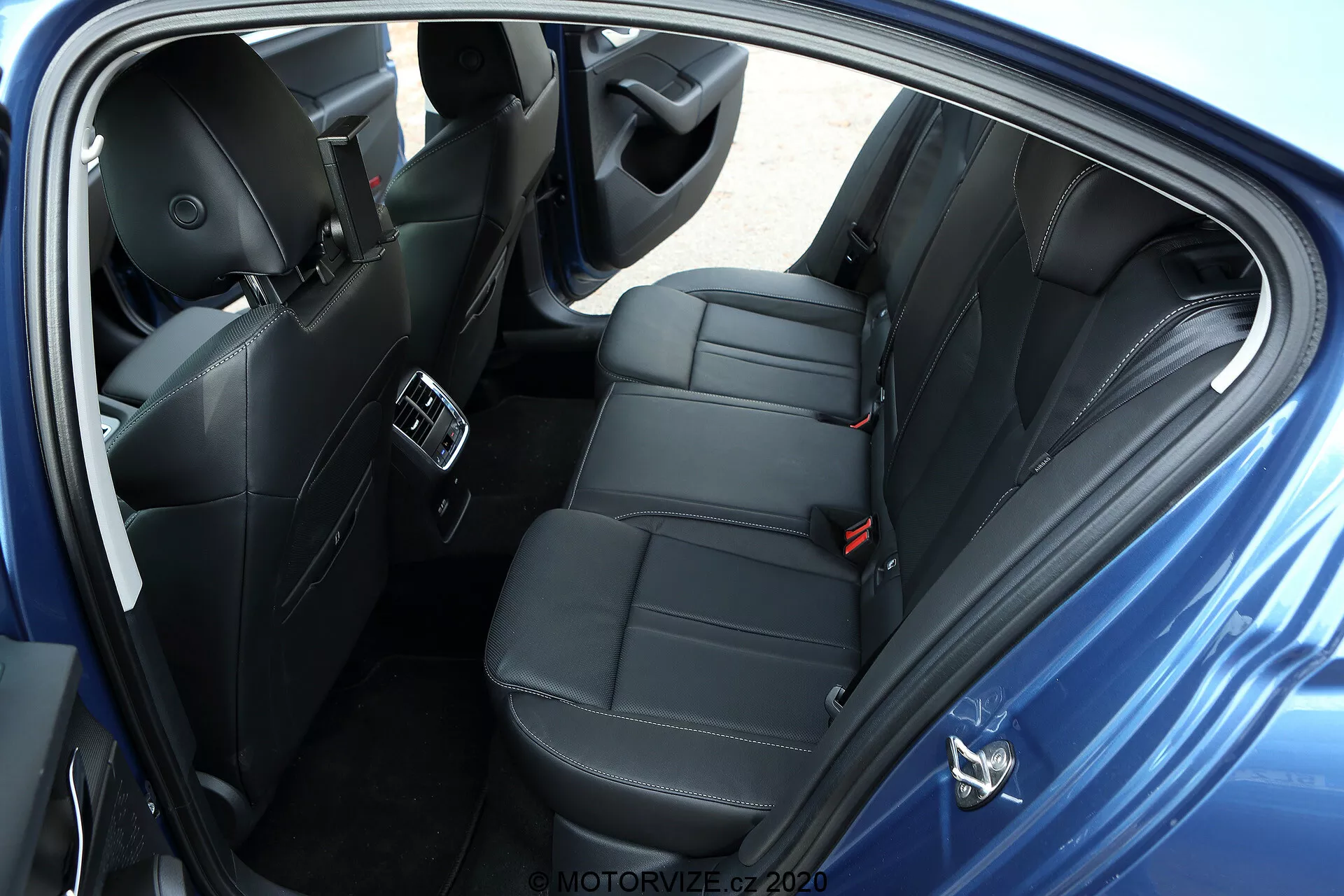 Felső nézetből készült kép egy Skoda Octavia 2020 limuzin hátsó utasteréről, a jobb oldalról. A fotón nyitott ajtóval láthatóak a modern, fekete bőr vagy bőrhatású ülések luxusvarrással. A hátsó üléspad két utas kényelmes elhelyezésére készült egyéni fejtámlákkal, ami sportos vagy kényelmi felszereltségre utal. A képen láthatóak az utasok hátsó részére telepített légkondicionáló szellőzők a középkonzolon, USB portok eszközök töltésére, fejlett hangrendszer hangszórói az ajtó paneljén, elektromos ablakvezérlők, tisztán látható biztonsági övek, beleértve a hátsó középső ülést rögzítő hárompontos övet is, valamint szőnyegpadlók. A sötét belső színséma egy prémium kategóriás, jól felszerelt járműre utal személyes tárgyak nélkül, parkolási helyzetben, semleges háttérrel.