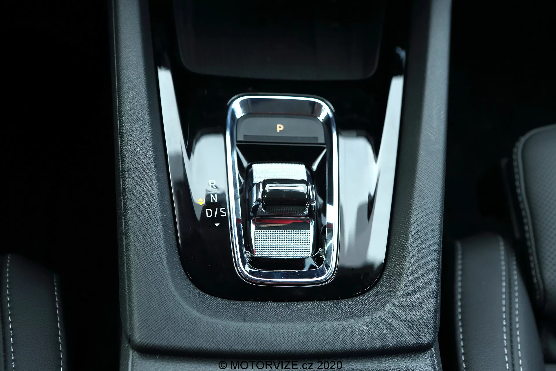 Közelkép egy modern váltóról a 2020-as Skoda Octavia Limousine középkonzoljában. A váltó szemből nézve elegáns, téglalap alakú, lekerekített élekkel és króm szegéllyel rendelkezik, a sebességfokozatok 'P', 'N', 'D/S' pozíciói láthatóak. Magas minőségű fekete anyagok veszik körül, látható varrással és egy finom hálós textúrájú ezüst felülettel, ami a luxust és részletek iránti figyelmet sugallja. Az erős világítás megvilágítja a robusztus, összetartó belső teret, ami fejlett elektronikus funkciókra és modern autóipari technológiákra utal.