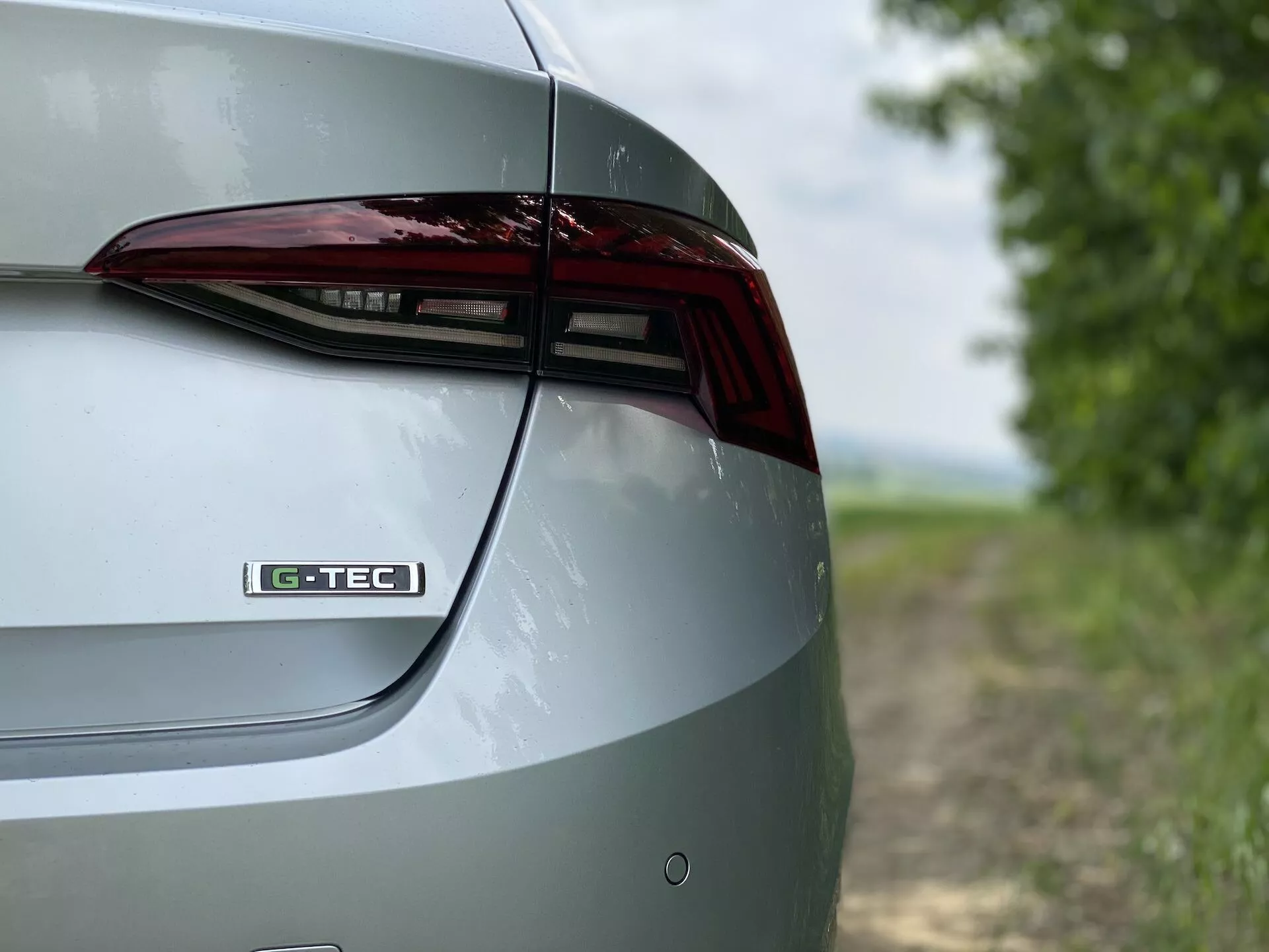 A Skoda Octavia 2020 limuzin jobb hátsó részének közeli, részletes nézete. A felvétel a csomagtartó fedelének, a LED-es hátsó lámpának és a hátsó lökhárító egy részének tervezési elemeit ábrázolja egy ferde, kissé megemelt szögből. A csomagtartó fedelén látható 'G-TEC' jelzés egy speciális technológiára vagy kivitelre utal. A fém festés kiemeli az autó polírozott külsejét. A homályos háttér vidéki környezetet teremt, amely kiemeli a jármű tulajdonságait és kialakítását.