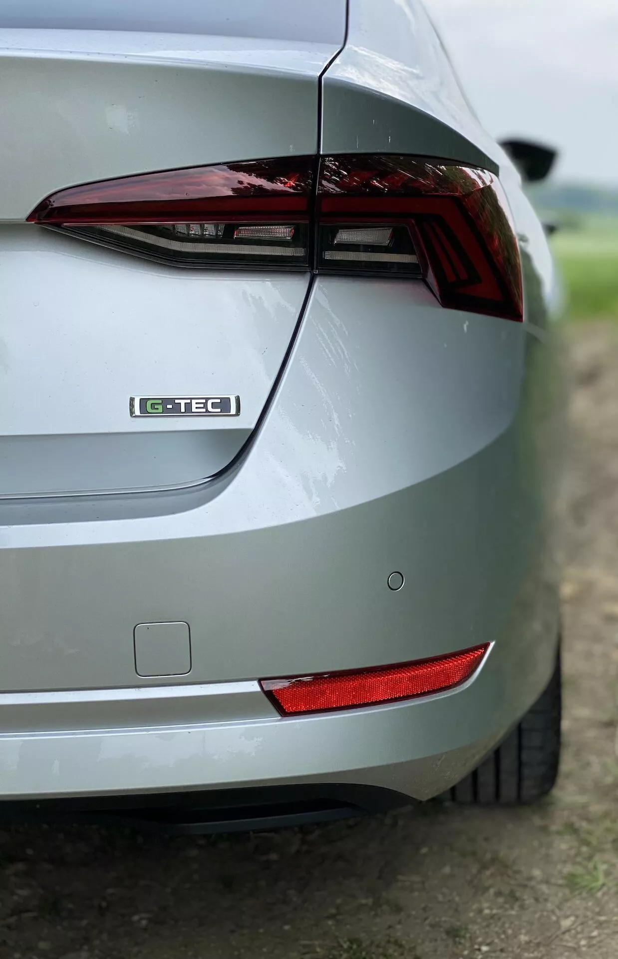 Közelképe egy ezüst Skoda Octavia Limousine 2020 jobb hátsó részének ferde nézetben. A képen látható a hátsó lámpa, a G-TEC jelvény, az elegáns csomagtér dizájn, a hátsó lökhárító, a hátsó reflektor és a metálfényes fényezés. A homályos háttér kiemeli az autó dizájnjának jellemzőit.