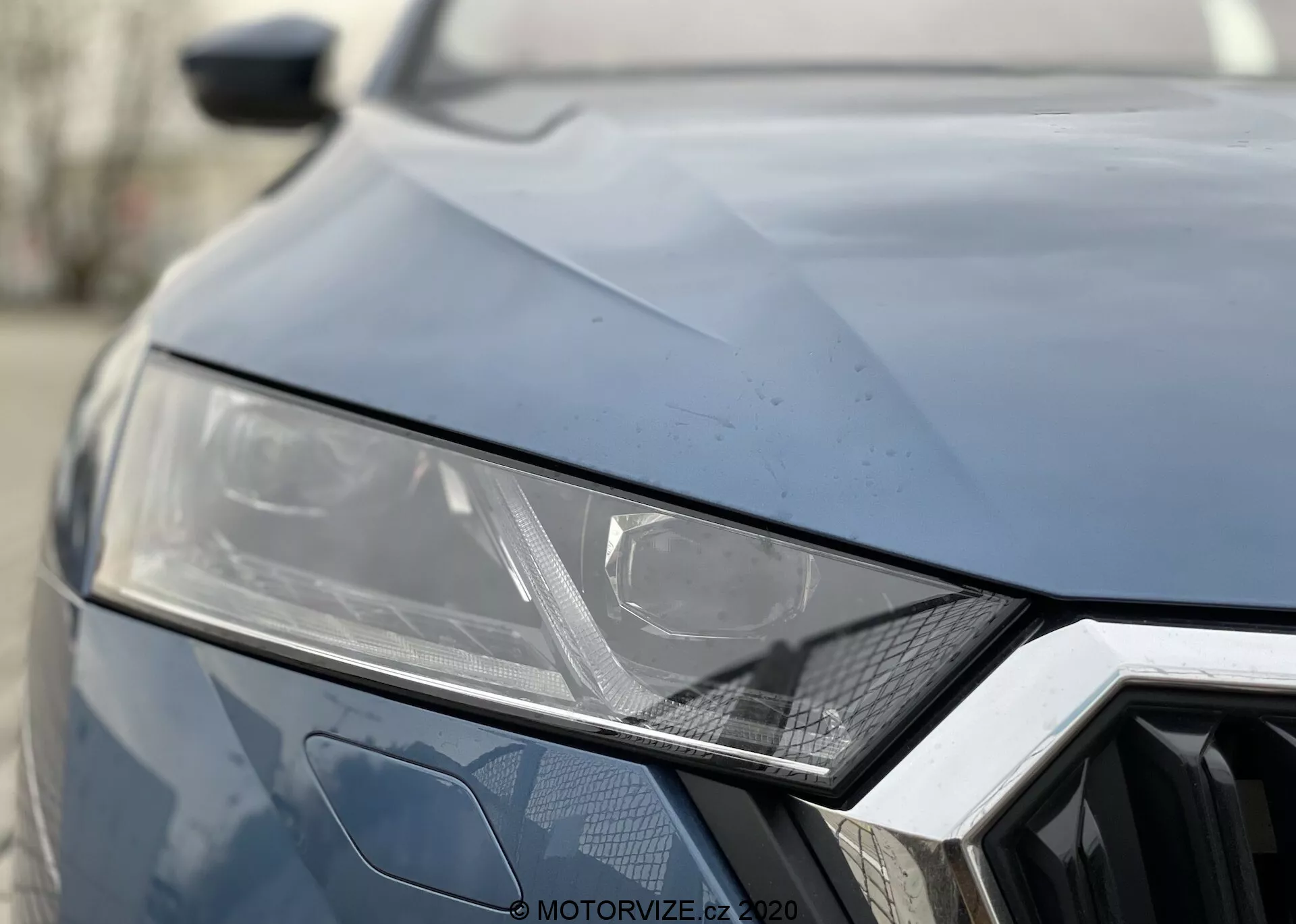 A Skoda Octavia 2020-es modell elejének közeli nézete. Sötét szín, mely az elegáns, szögletes kialakítású bal első lámpát mutatja, amely LED vagy HID technológiát sugall, geometrikus hatszög mintás hűtőrács és szögletes gyűrődések a motorháztetőn sportos megjelenés érdekében.