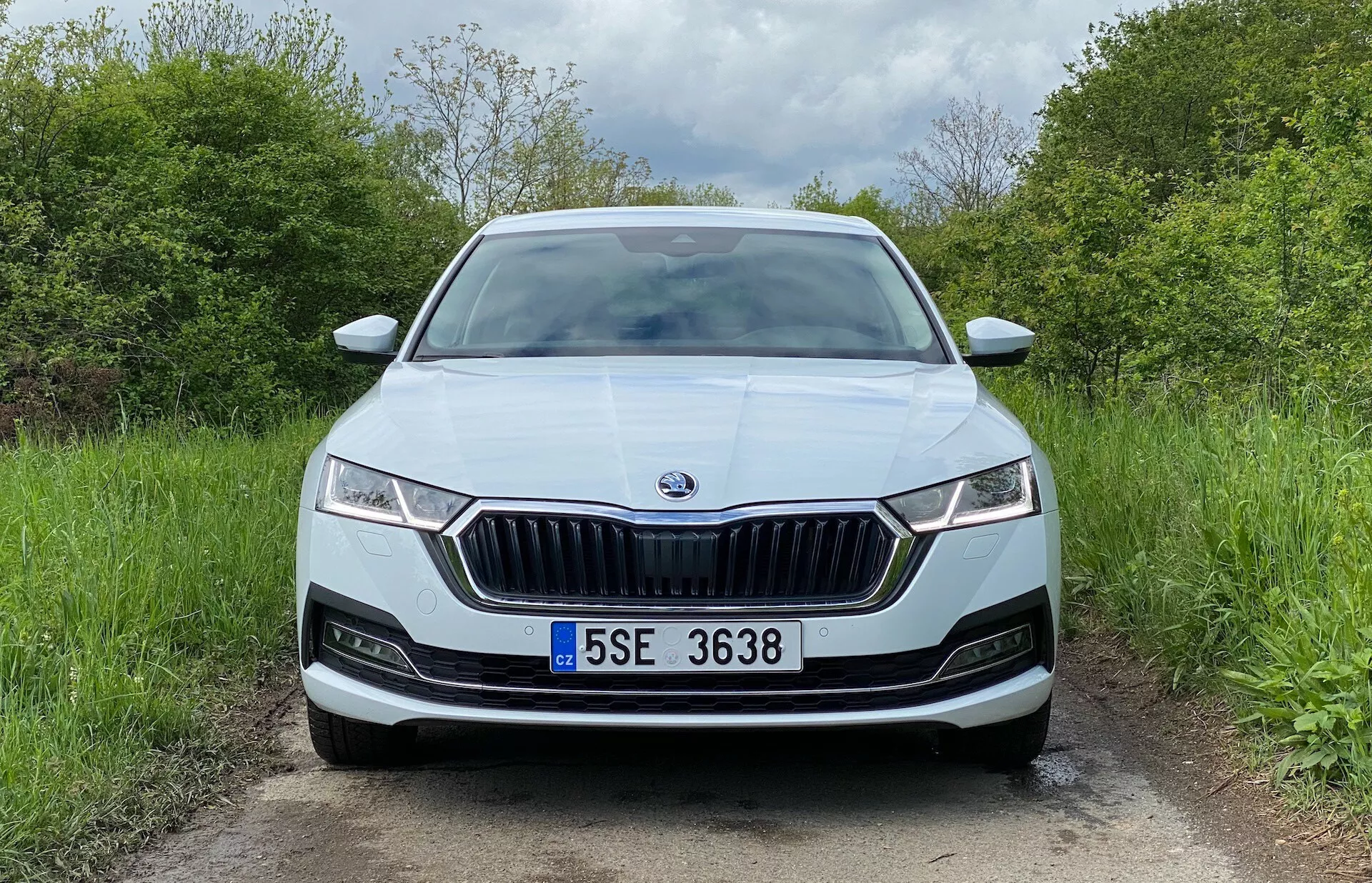 A Skoda Octavia 2020-as szedán elölről egy vidéki úton, kiemelve a jellegzetes, függőleges lécekkel és krómkerettel ellátott hűtőmaszkot, keskeny LED fényszórókat DRL-ekkel, központi márkaemblémát, a lökhárítóban található parkolószenzorokat, középen elhelyezett rendszámtáblát, fémes festést, légbeömlő rostélyokat és esetleges ködlámpákat.