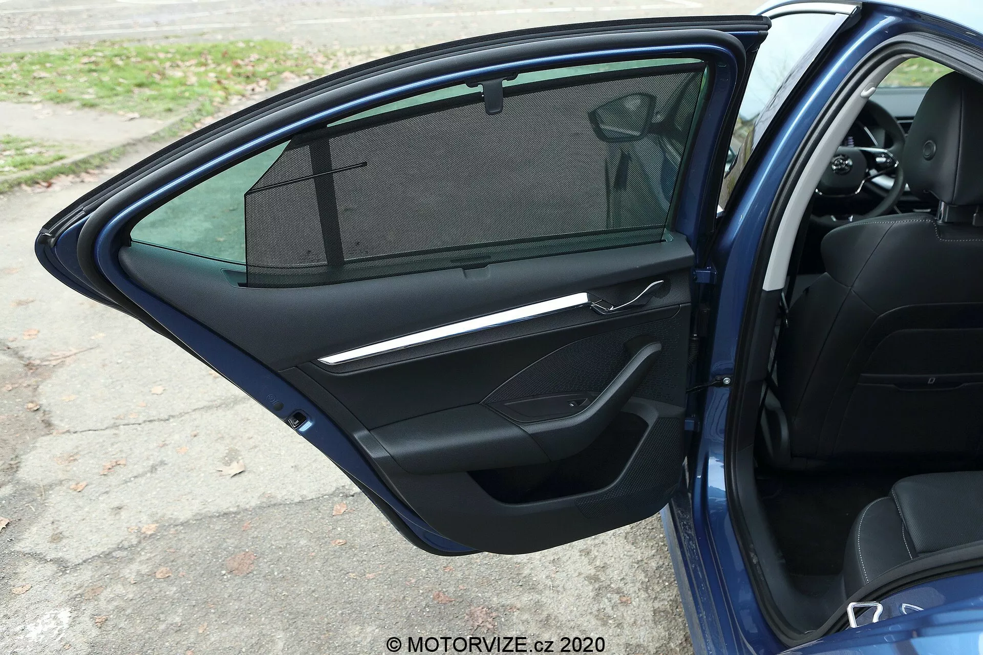 Közelkép a Skoda Octavia 2020 limousine nyitott első utasajtójáról, amely egy sötét színű belső ajtópanelet mutat, fémbetétes ajtókilinccsel, ablakemelő kapcsolókkal, tárolórekesszel, hangszórórácssal, részben látható küszöbbel és napellenzős ablakkal.