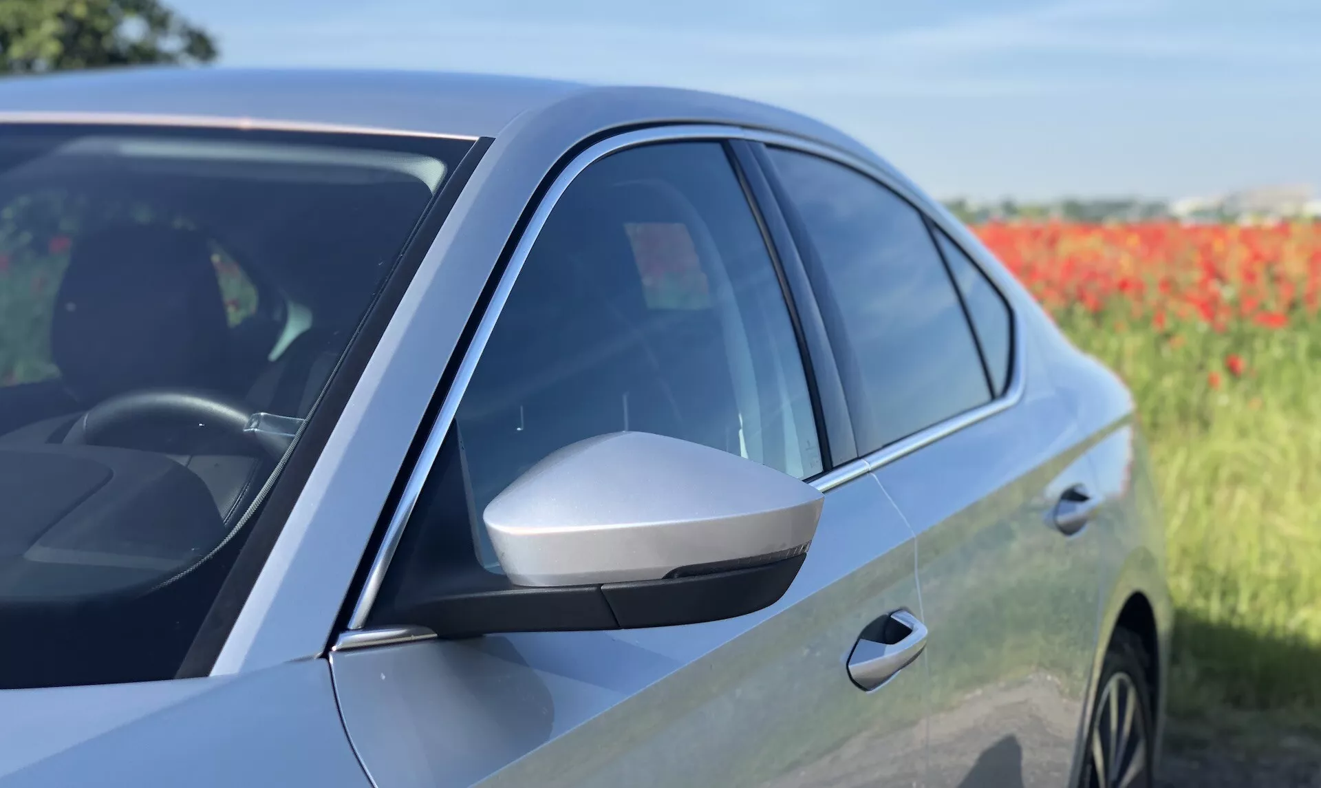 Közelről oldalnézetben egy modern Škoda Octavia 2020 szedán, amely a vezető oldali első ajtót és az oldalsó tükröt mutatja. A kép kiemeli a karcsú, integrált irányjelzővel ellátott oldalsó tükröt, a sötétített ablakokat, a fényes festékfelületet és az aerodinamikai dizájnt középtől az eleje felé. Az ajtópanel és az első kerékjárat részének tükröződése az ötvözet felnik jelenlétét sugallja. A fotó haladó tervezési jellemzőkre és a jármű potenciális technológiáira hívja fel a figyelmet egy kissé elmosódott természetes háttér előtt.