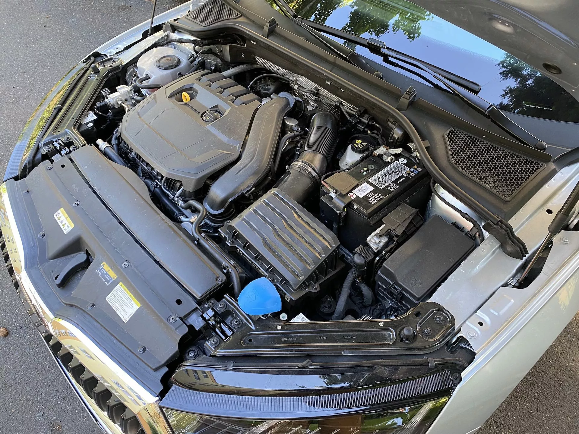 Közelkép a Skoda Octavia 2020 limuzin motorteréről nyitott motorháztetővel, különféle alkatrészeket bemutatva, beleértve a középre helyezett motorburkolatot, a jobb oldalon akkumulátort, a bal oldalon hűtőbordákkal ellátott téglalap alakú komponenst és egy folyadéktartályt kék kupakkal. A lejtős, kissé oldalirányú szögből készített kép a szélvédő alján elhelyezkedő fekete műanyag borítást is megmutatja, kiemelve a gépjármű motorterének technikai bonyolultságát.