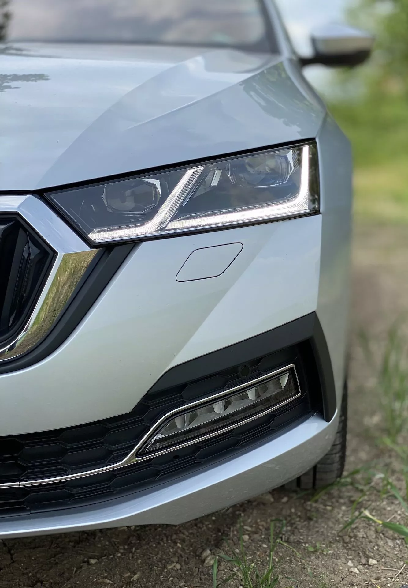 Közelkép a Skoda Octavia 2020 (Octavia Limousine) elülső baloldali részéről, a hűtőrács közepétől a bal fényszóróig, a motorháztető egy részéig, valamint az első lökhárító és a sárvédő egy részéig mutatva a részleteket. Az alacsony szögből készült kép kiemeli az elegáns fényszóró dizájnt LED-es nappali menetfényekkel, króm díszítésű hűtőrácsot és az alsó rács modern, fémes kivitelét. Az élesség az autó részleteire fókuszál, kis mélységélességgel, amely professzionális, magas minőségű megjelenést eredményez.
