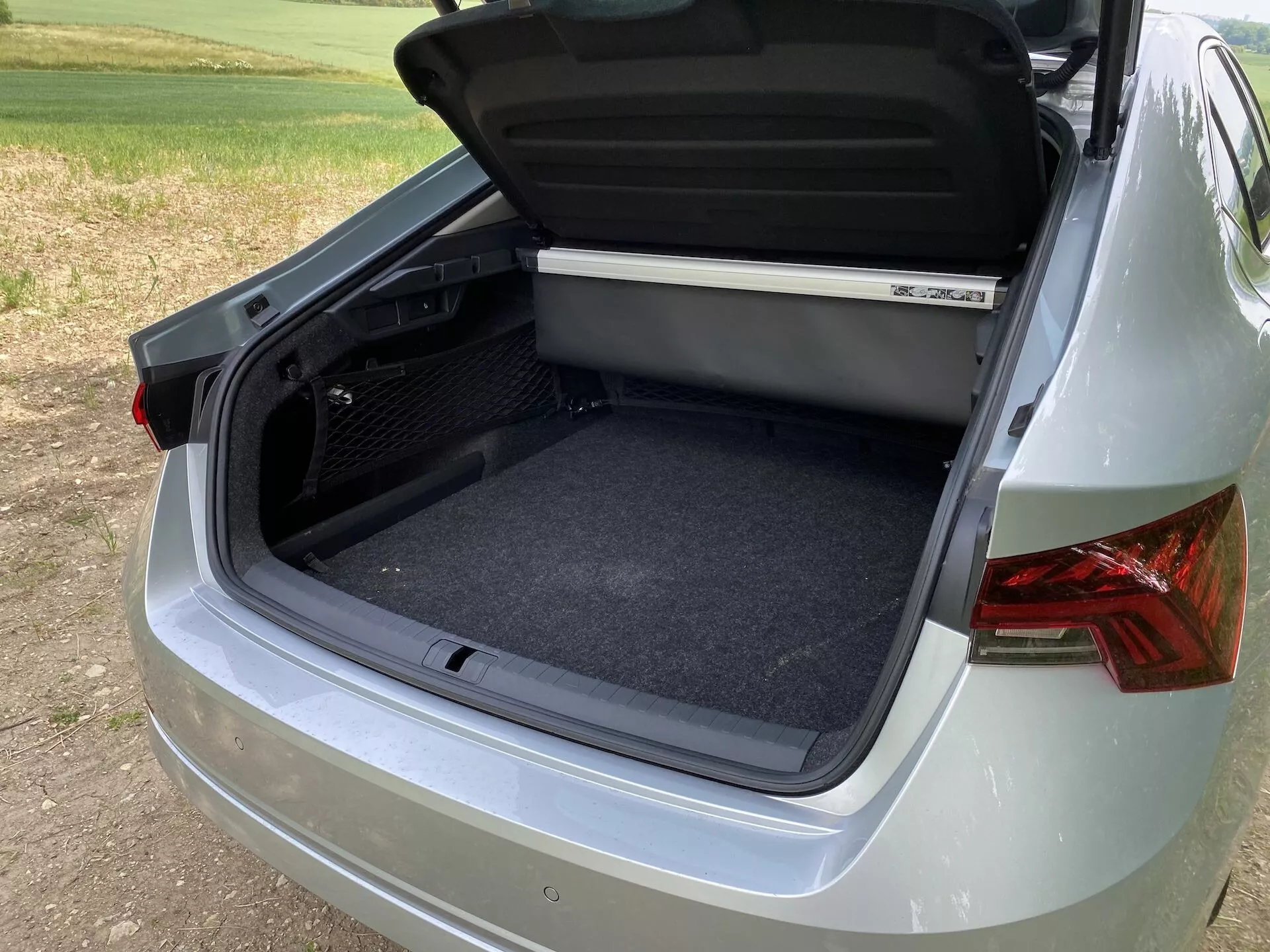 Közeli háromnegyedes nézet egy Škoda Octavia 2020 hátsó csomagtér béléséről, amely az üres, tiszta, szőnyeggel borított belső térre összpontosít. A szög a jármű jobb oldalának és hátuljának egy részét is megmutatja, modern designú hátsó lámpákkal, sima, metálfényezéssel és részben látható lökhárítóval. A csomagtérfedél alján ergonomikus fogantyú található, és a csomagtér tetején egy polc is látható. A környezet külső tér, puha nappali fényviszonyokkal.