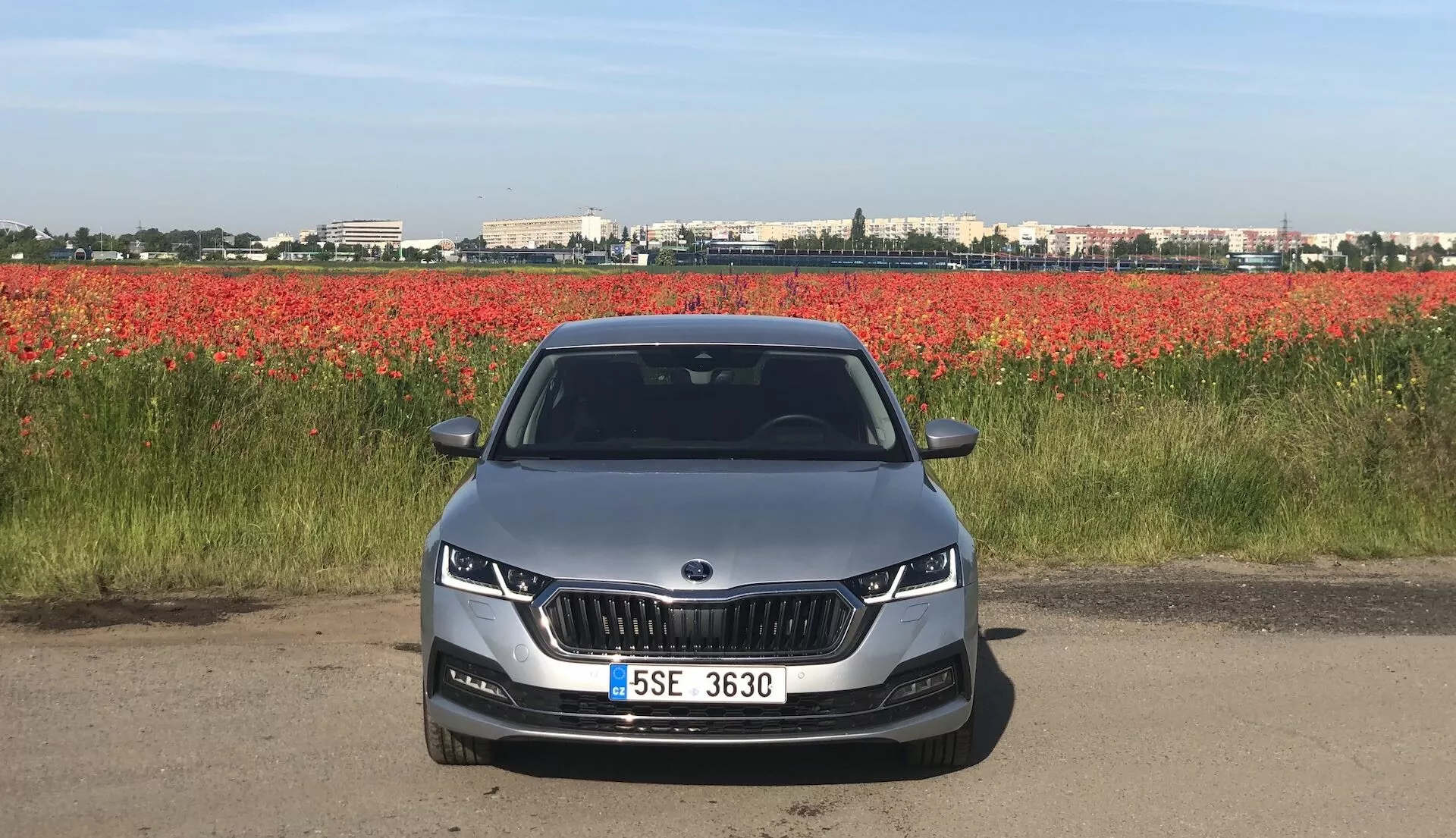 Egy 2020-as Skoda Octavia Limousine elölnézete egy úton vagy földúton, mely előtérbe helyezve mutatja meg a jellegzetes veseshéj alakú hűtőrácsot és az elegáns LED vagy adaptív fényszórókat. Az aerodinamikus első lökhárító ködlámpa-kivágásokat és légbeömlőnyílásokat tartalmaz. A motorháztető vonalainak kontúrjai esztétikai és aerodinamikai dizájnt sugallnak. A teljesen látható szélvédő fejlett technológiai funkciókra, például head-up kijelzőkre vagy vezetőtámogató rendszerekre utalhat. Világos színű autó, középen elhelyezett rendszámtáblával és parkolószenzorokkal, vörös virágok hátterében.