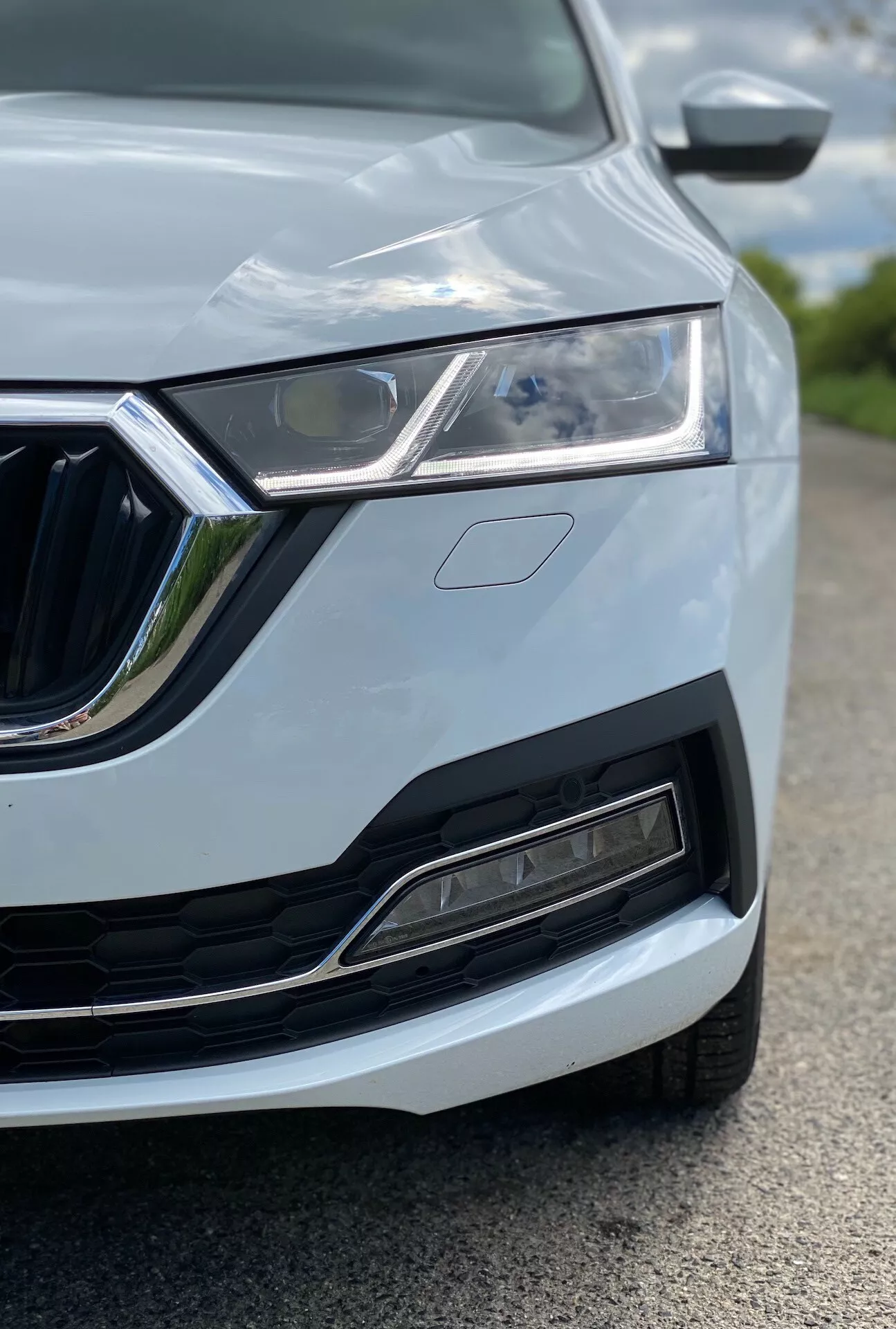 Közelkép egy Skoda Octavia 2020 (octavia-limousine) bal első negyedéről, kiemelve a LED-es fényszórót, a jellegzetes, krómozott részletekkel ellátott hűtőrácsot, a modern első lökhárítót légbeömlővel és érzékelőkkel, a gyűrődéses motorháztető dizájnt és a részben látható oldal tükröt integrált irányjelzőkkel. Az autót világos színűre festették, valószínűleg fehérre vagy ezüstre, elegáns karosszéria vonalakkal. A kép alacsony perspektívából készült, a fényszóróra és a hűtőrácsra koncentrálva, sekély mélységélességgel, ami elmosódássá teszi a környező elemeket.