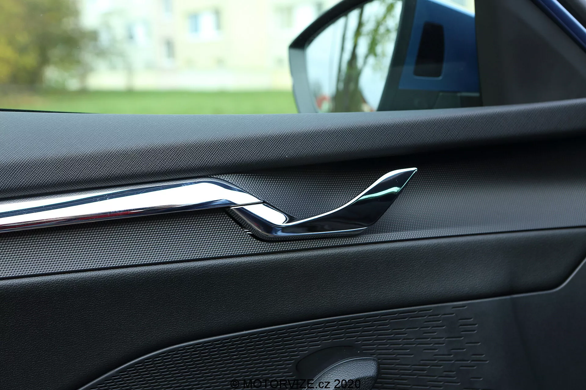 Közelkép egy 2020-as Skoda Octavia belső teréről, amely az első utasajtó hátsó részére fókuszál, kiemelve a tükröződő krómozott ajtókilincset. Az ajtópanel bőr looking vagy texturált puha tapintású anyaggal van bevonva, látható varrásokkal, ami magas szintű belső befejezésre utal. Jól látható egy hangszórórács is, amely az autó audio rendszerének része, egységes mintázattal és színkoordinált kialakítással. A kép a hátsó ülésből készült, ami arra utal, hogy az autó egy lakóhelyi területen parkol, lágyan elmosódó zöld háttérrel.