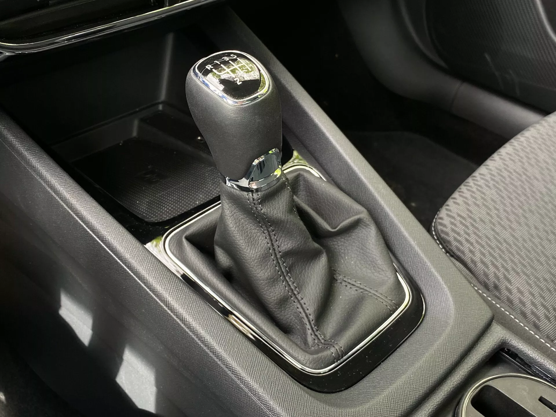 Madártávlatú közelkép a Skoda Octavia 2020 (Octavia-Limousine) manuális sebességváltójáról. A fekete bőrből készült váltógomb ezüst kiemeléssel mutatja a hatsebességes váltómintázatot és a hátramenetet. A váltógomb talapzata, amelyet ezüst keret szegélyez, fekete bőrből készült burkolatban található. A közelben tárolórekesz, középkonzol és pohártartók láthatók, mindezt nappali fény világítja meg.