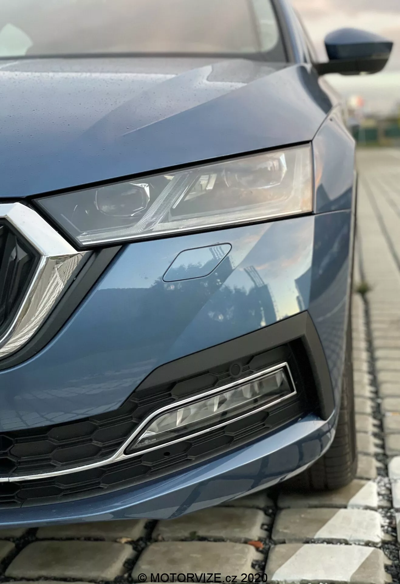 Közeli részletkép egy modern 2020-as Skoda Octavia-ról alacsony szögből fényképezve, amely a bal elülső részre összpontosít, beleértve a motorháztetőt, elegáns LED fényszórót, geometrikus hűtőrácsot és az első lökhárítót légbeömlővel. Fémes kék festés, szögletes kialakítási elemek és kiemelt aerodinamikai jellemzők.
