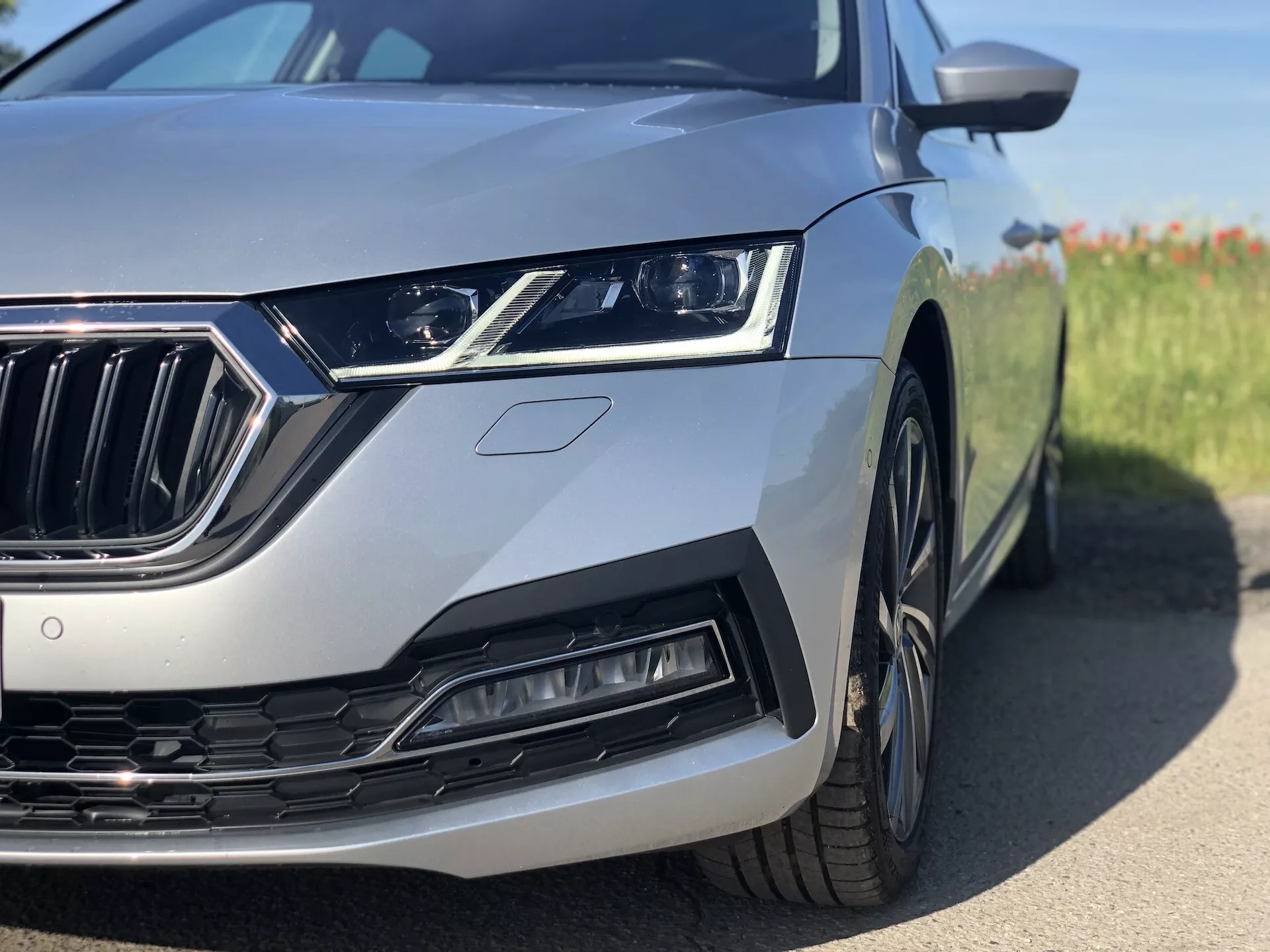 2020-as ezüst Skoda Octavia limuzin bal első negyedének közeli képe, kiemelve a karcsú LED-es fényszórót, a függőleges lamellákkal ellátott krómozott hűtőrácsot, a formázott légbeömlőt és a részben látható kereket és sárvédőt. Az alacsony, talajszintű szögből készült felvétel a modern formatervezési elemekre és a fejlett technológiai jellemzőkre összpontosít, elmosódott háttérrel.
