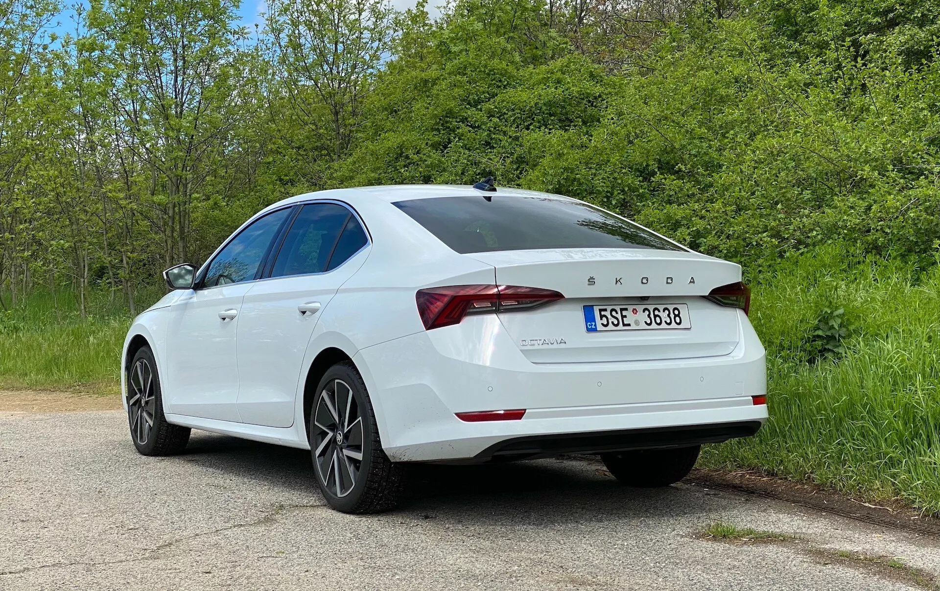 A kép egy fehér, 2020-as Škoda Octavia szedánt mutat be, amely az autó jobb oldalát és hátsó alkatrészeit kiemelő hátsó háromnegyed szögből készült. A főbb részletek közé tartoznak a LED-es hátsó lámpák, a csomagtérajtó közepén található Škoda embléma és az alatta feltüntetett OCTAVIA felirat. Az TDI felirat a csomagtérajtó jobb oldalán jelzi a valószínűleg turbófeltöltős közvetlen befecskendezésű dízelmotort. A csomagtérajtó formás kialakítással rendelkezik, amely kissé megemelkedő ajak spoilerként is funkcionálhat, ami fokozza az autó esztétikáját és aerodinamikáját. Az autó ötküllős alufelnikkel van felszerelve, amelyek teljesítmény és komfort közötti egyensúlyt sugallhatnak. Az oldalnézet egy simán lejtő tetővonalat tár fel, amely a modern szedánokra jellemző, és javíthatja az aerodinamikát és sportos megjelenést. A hátsó lökhárító látható, beépített parkolószenzorokkal, amelyek az autó technológiai képességeit sejtetik. A festék csillogó és jól karbantartott, a karosszéria nem mutat sérülés jeleit. Az autó egy nyugodt, egyirányú vidéki vagy parkúton parkol, amelyet mindkét oldalon növényzet szegélyez. Nincs közvetlen közelségben más jármű, a környezet nyugodt és zavartalan. Ez a konkrét kép alkalmas a külső dizájnelemek bemutatására, és hasznos lehet az autótervezéssel, a modern szedánokkal, az autótechnikával kapcsolatos kategóriákban, különösen, ha figyelembe vesszük a dízelmotort és a vezetéstámogató funkciókat, mint például a parkolószenzorokat. A fotó kompozíciója, az autó körüli elegendő térrel, lehetővé teszi, hogy a néző kizárólag az autóra koncentráljon.