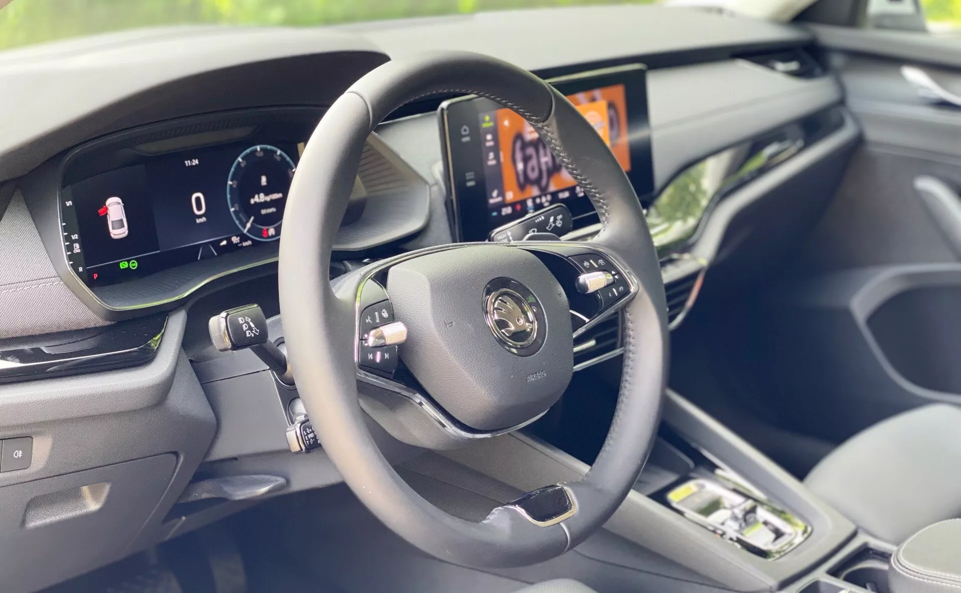 Modern jármű belső tere, amely a vezetőoldali műszerfalra összpontosít a vezetőüléshez képest enyhén jobbról nézve. A képen központi szerepben egy háromküllős kormánykerék található vezérlőgombokkal, egy teljesen digitális műszercsoport, amely a jármű sebességét és egyéb fontos információkat jeleníti meg, és egy nagy felbontású központi infotainment kijelző, amely az autó klímaszabályozó rendszerét mutatja. Az elegáns, puha tapintású anyagok és a műszerfalon valamint az ajtón látható díszítések felerősítik az autó belsejének prémium érzését. Az elmosódott előtér egy utasülés meglétét sejteti.