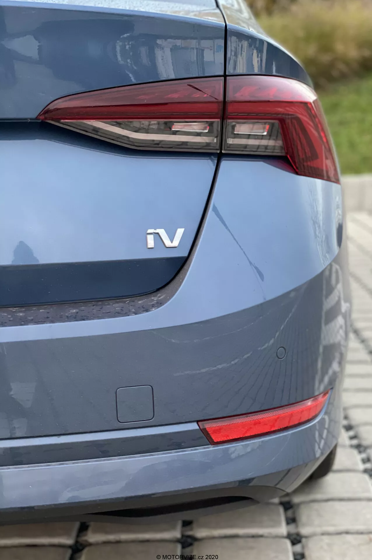 Közelkép egy 2020-as Skoda Octavia hátsó részéről ferde szögből, középpontban a modern, szögletes hátsó lámpával, valószínűleg LED elemekkel. A hátsó lámpa alatt egy kis téglalap alakú fényvisszaverő vagy kiegészítő lámpa látható. A kékesszürke fémes karosszéria ívelt csomagtérajtót, jól integrált hátsó lökhárítót és egy hybrid vagy elektromos hajtásláncot jelző „iV” feliratot tartalmaz. A jobb alsó sarokban alig látható parkolószenzorok fejlett vezetőtámogató rendszerekre utalnak. A nappali fényviszonyok mellett, kis mélységélességgel készült fénykép egy macskaköves felületen parkoló autót ábrázol.