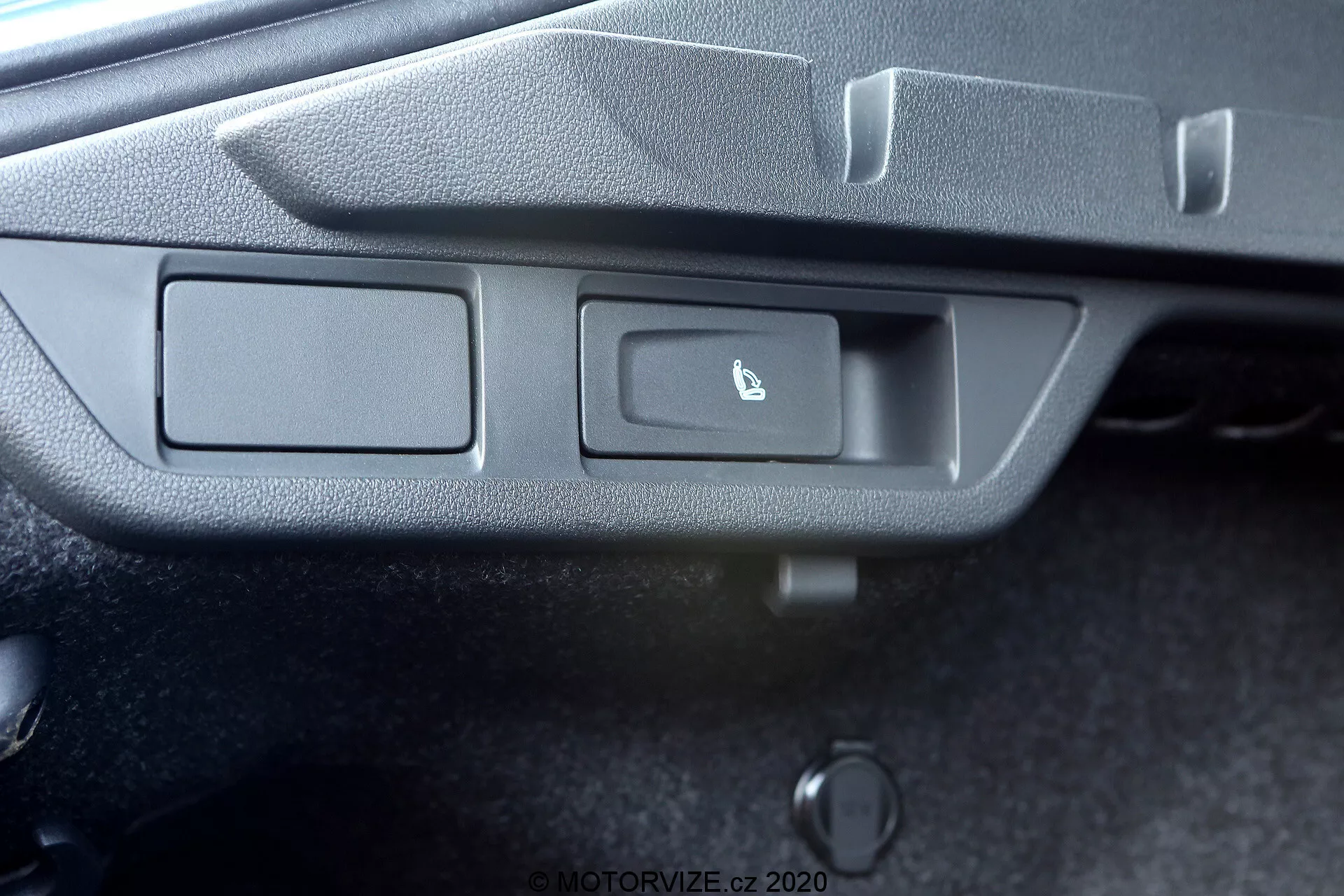 A képen egy autó vezérlőpaneljének részletes nézete látható enyhén szögletes felülnézeti perspektívából. A panel két téglalap alakú gombot tartalmaz, amelyek egy texturált szürke műanyag keretbe vannak beépítve. A bal gomb jelöletlen, míg a jobb gomb egy félig kinyitott csomagtérajtó ikonját viseli, jelezve a csomagtér nyitási funkcióját. Egy kis világítós jelzőfény, valószínűleg LED, helyezkedik el a gombok jobb oldalán. A gombok alatt van egy hengeres tárgy, feltehetően kézi vagy vészhelyzeti csomagtér nyitó készülék. A fotó nem tárja fel az autó belső részének szélesebb kontextusát.