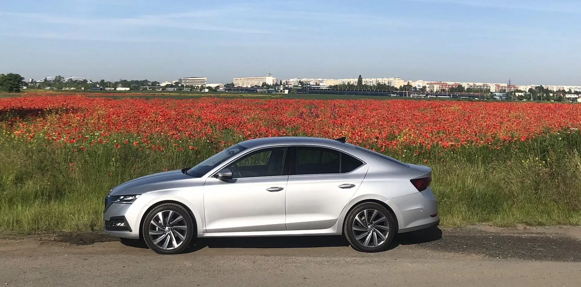 Egy modern, ezüst színű Skoda Octavia 2020 szedán oldalnézete egy sík felületen, piros virágok mezője mellett parkolva, elegáns aerodinamikus dizájnnal, LED fényszórókkal, integrált irányjelzőkkel ellátott karosszéria színű oldalsó tükrökkel, alufelnikkel, négy ajtóval, sötétített ablakokkal és finoman lejtő tetővonallal.