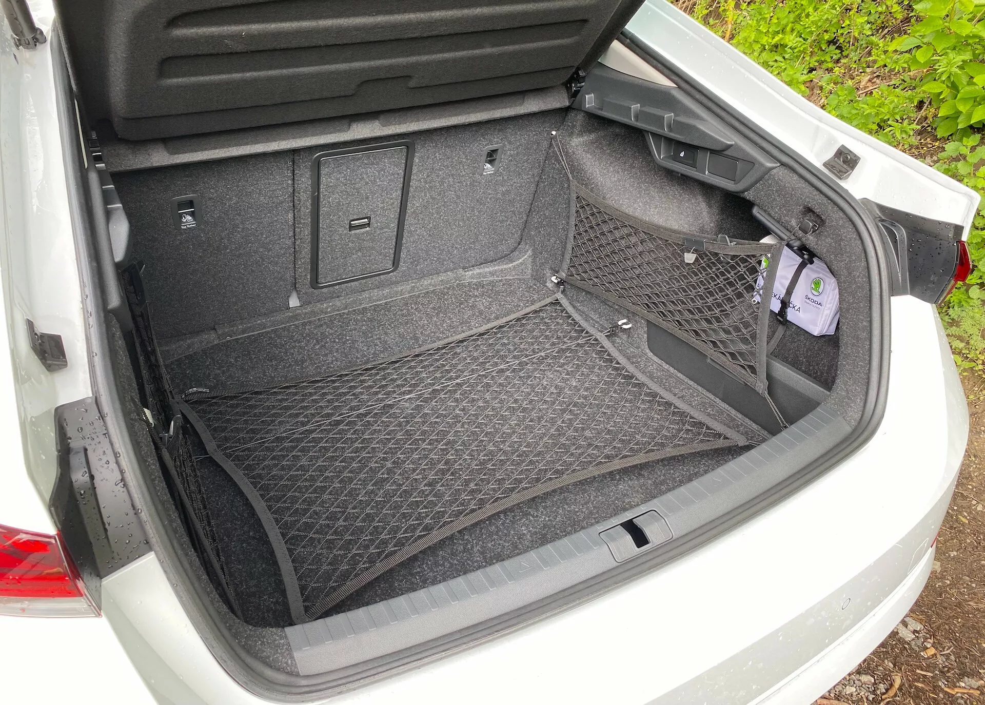 Hátulnézetből nyitott Škoda Octavia 2020 (octavia-limousine) csomagtér szőnyegezett felülettel és rakományrögzítő hálós tartozékkal. Jellemzői közé tartozik a műanyag burkolat a védelemért, különböző kampók és rögzítési pontok további rakomány opciókhoz. A jobb oldalon egy kis, fedővel ellátott rekesz található, amely további tárolást vagy alapvető szerszámokhoz való hozzáférést biztosít.