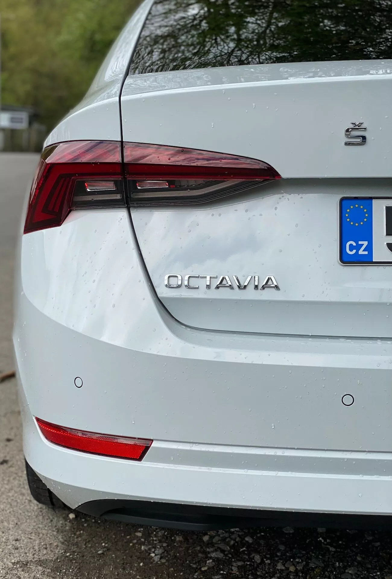 Részletes közelkép egy 2020-as fehér Škoda Octavia hátsó részéről, háromnegyedes perspektívában, a jármű jobb oldalára fókuszálva. Az kép kiemeli az olyan fontos jellemzőket, mint a szögletes LED hátsó lámpa, a hátsó lökhárító reflektív elemei, az 'OCTAVIA' név és a Škoda logó a csomagtartófedélen, valamint részletezett európai uniós rendszámtábla 'CZ' megjelöléssel. Finom vízcseppek a felületen azt jelzik, hogy a járműt nemrégiben eső érte vagy lemosták, ami növeli a frissességet a fehér festésen, miközben enyhe szennyeződést mutat a rendszámtábla körül.
