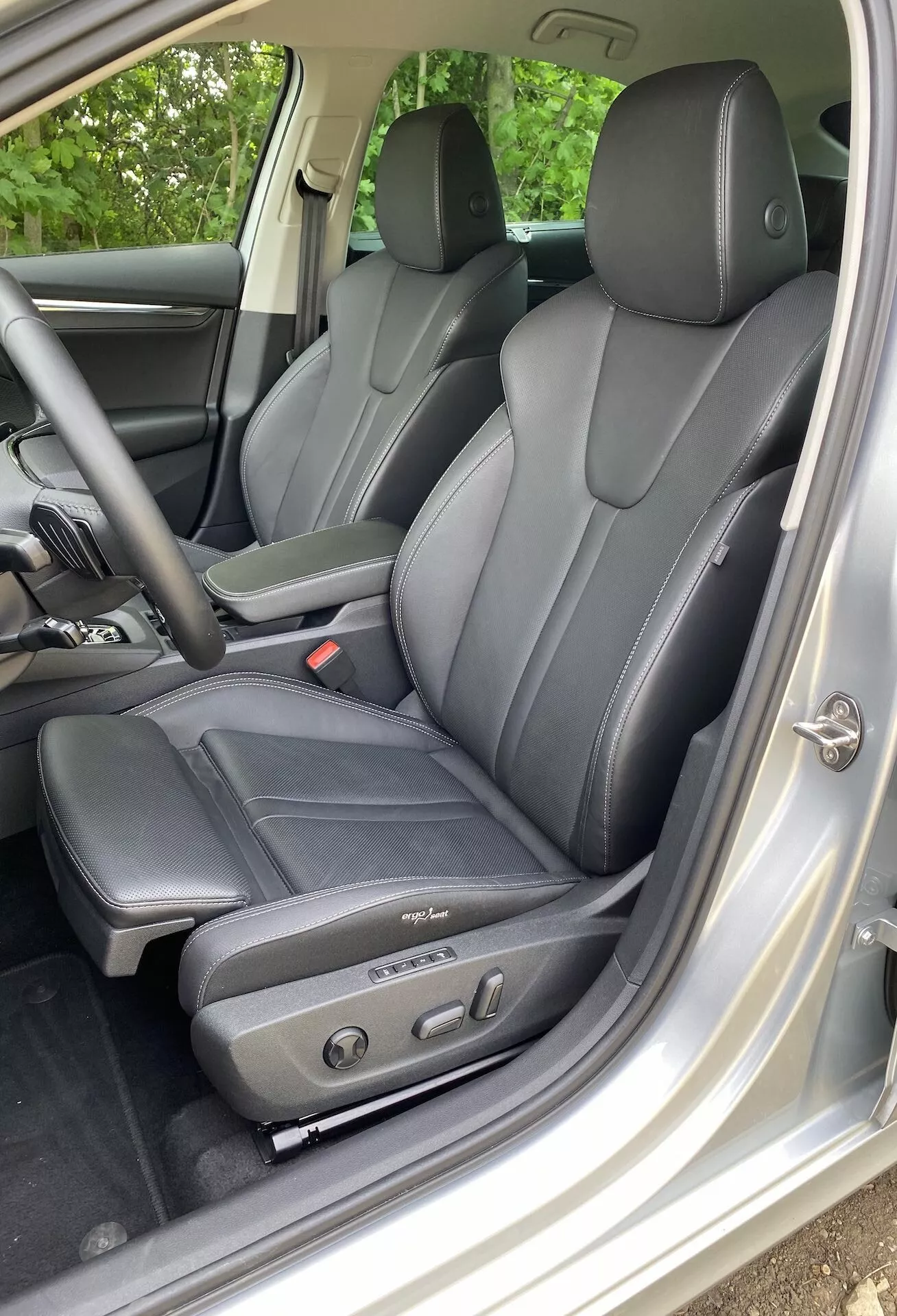 A 2020-as Škoda Octavia limuzin belső nézete a sofőr részére összpontosítva, sötét bőrülésekkel, ergonomikus kialakítással és elektromos beállítási vezérlőkkel, bal oldalról, a nyitott vezetőoldali ajtón keresztül nézve. A látható fejtámlák a sofőr és a hátsó utasok számára legalább négy utas befogadását jelzik. Az ezüst vagy világosszürke belső dekoráció kontrasztot képez az ülésekkel, az ajtópaneleken található ablakszabályzó és tükörvezérlő kiemeli a technológiai kényelmet és luxust.