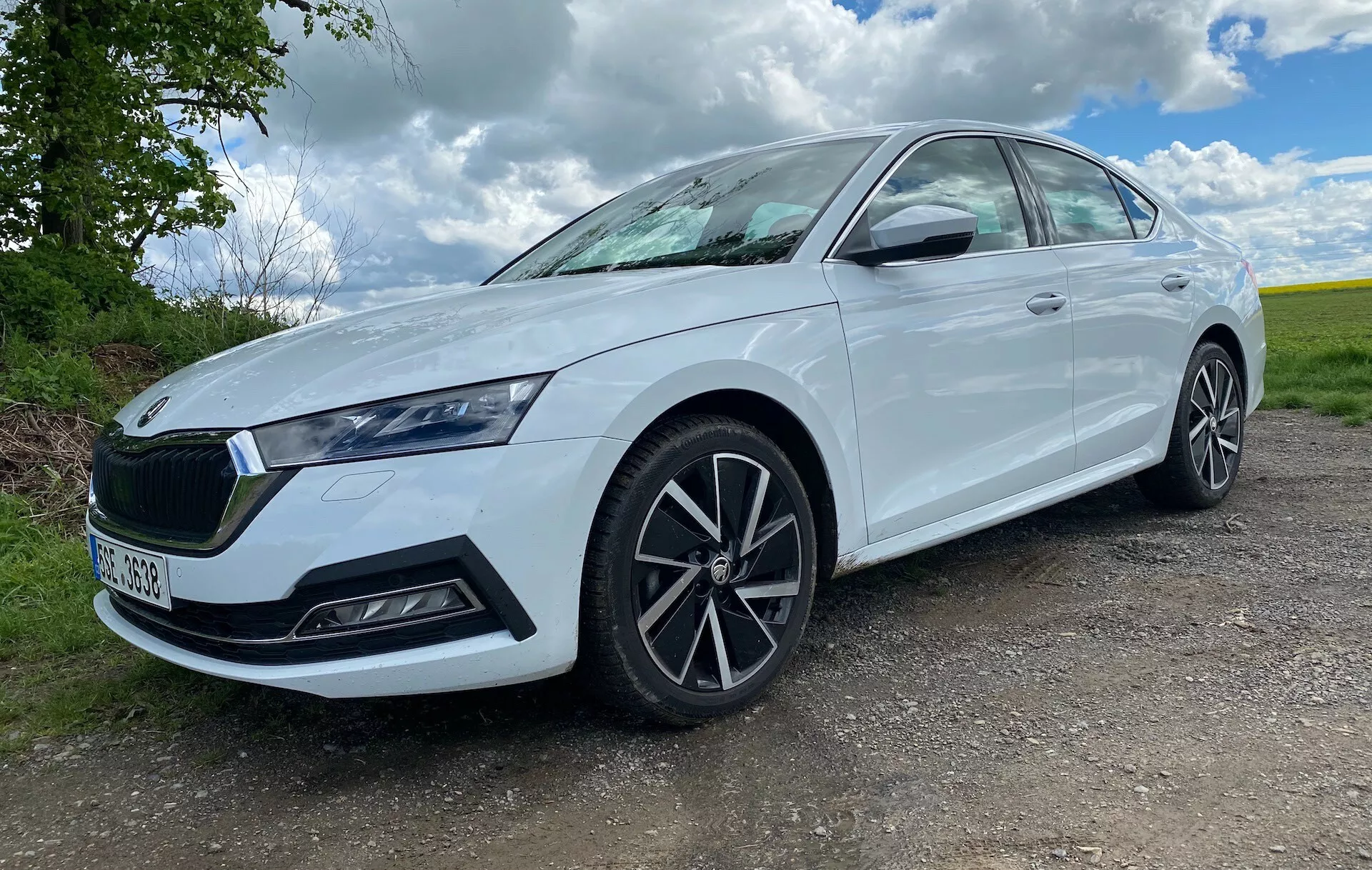 Ez a kép egy 2020-as Skoda Octavia limuzint ábrázol alacsony, háromnegyedes elölnézetből, kiemelve a hűtőrácsát, fényszóróit és az első kerék dizájnját. Földszintről készítve a fotó hangsúlyozza az autó állását, impozánsabbá téve azt. Az autó bal oldalát mutatja, enyhén elölről szögelve, ezáltal látszik az autó orrésze és oldalprofilja. A fehér szedánt nappali fényben fényképezték egy vidéki háttérrel, növényzet és részben felhős égbolt mellett délben. A karcsú fényszórók LED nappali menetfényekkel, a jellegzetes hűtőrács márkaemblémával, a többlábas könnyűfém kerekek és a integrált irányjelzők az oldalsó tükrökön jól láthatók, ugyanúgy, mint az első lökhárítón lévő légbeömlők vagy biztonsági szenzorok. Az autó új vagy jól karbantartott állapotban van, enyhén sötétített ablakokkal és tiszta felülettel, ideális eladási hirdetésekhez vagy autós véleményekhez.