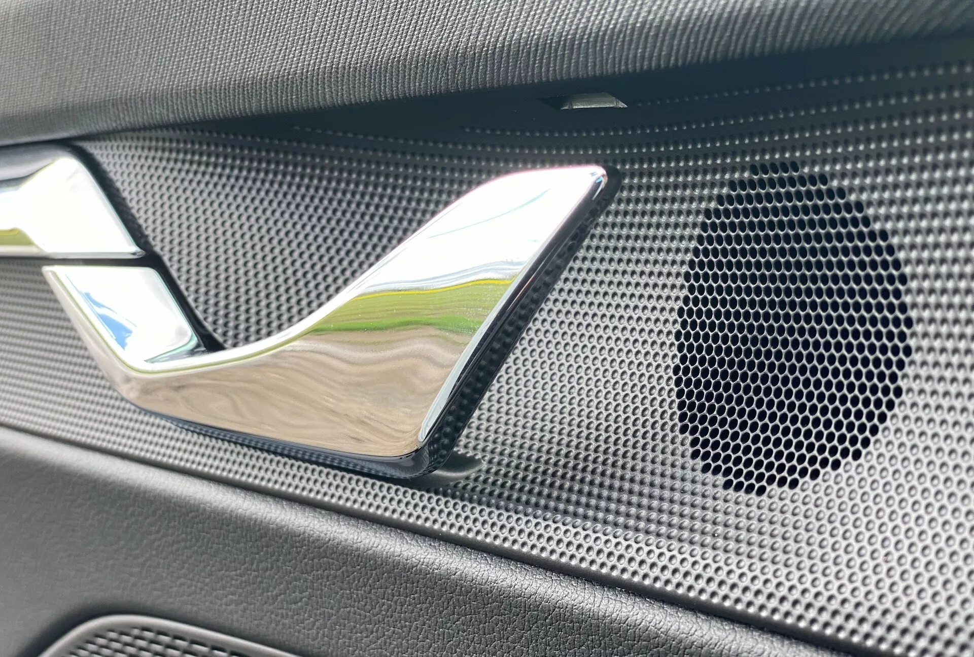 A 2020-as Skoda Octavia limuzin belső részletének közelije, kiemelve a krómozott ajtókilincset és a méhsejt mintás hangszórórácsot textúrált, esetleg karbon anyagból, hangsúlyozva a luxust és a csúcsminőségű audiorendszert.