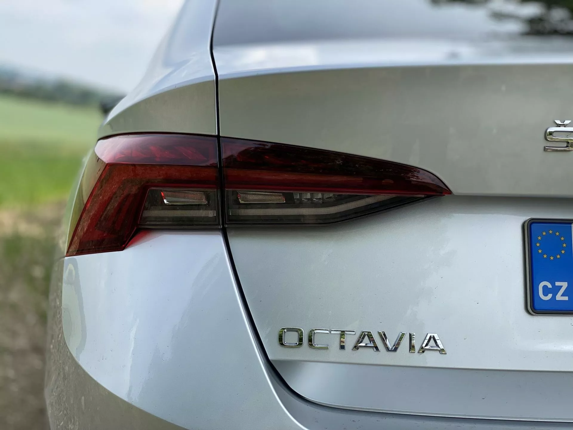 Közelkép egy ezüst színű 2020-as Škoda Octavia hátsó jobb oldaláról. A kép megörökíti a hátsó lámpák éles és szögletes kialakítását, beépített féklámpákkal, irányjelzőkkel és valószínűleg tolatólámpákkal. A csomagtartó fedelén feltűnően jelenik meg az 'OCTAVIA' modell megnevezés krómozott betűkkel a hátsó lámpa alatt. Egy kis kék és sárga csillagokkal ellátott téglalap jelzi a Cseh Köztársaságba való regisztrációt. A jármű polírozott, fémes fényezése tükrözi a környező tereket, melyet egy elmosódott növényzet hátterébe helyeztek, kiemelve annak kifinomult kialakítását.