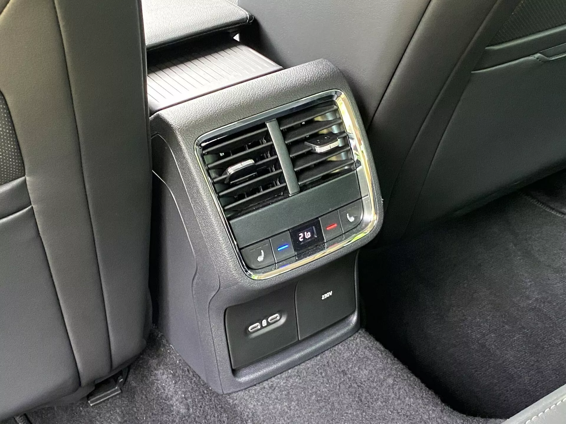 Részletes nézet a hátsó klímavezérlő konzolról egy 2020-as Skoda Octavia limuzin hátsó részében. Előre nem központosan rögzítve, jól látható a középen elhelyezkedő panel az első kartámasz hátulján. Digitális klímaszabályozó gombok hőmérséklet-beállítással, függőleges szellőzőnyílások, 21℃-ra beállított digitális hőmérséklet kijelző, USB töltőportok és a hátsó ülések fűtési vagy hűtési funkciójának lehetősége található meg rajta. Az anyagok műanyag és szövet kombinációja fekete és sötétszürke színben, természetes megvilágítás mellett.