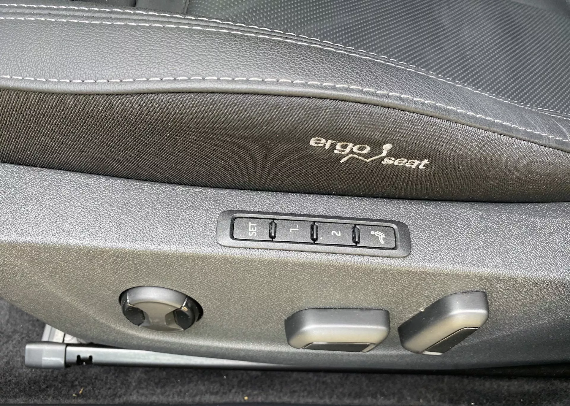 Közeli felvétel egy Škoda Octavia 2020 belteréről, az ergonomikus ülés állító gombjaira fókuszálva. Az ülés sötét felső részén 'Ergo seat' felirat van hímezve. A felirat alatt öt gomb látható: egy az ülés memória beállításához és négy az ülés előre, hátra, fel, le és háttámla dőlésszögének állításához. Továbbá, egy kerek tárcsa is található, ami valószínűleg a deréktámasz állítására vagy más többfunkciós ülésbeállításokra szolgál, jelezve a jármű magasabb kényelmét és fejlett technológiáját.