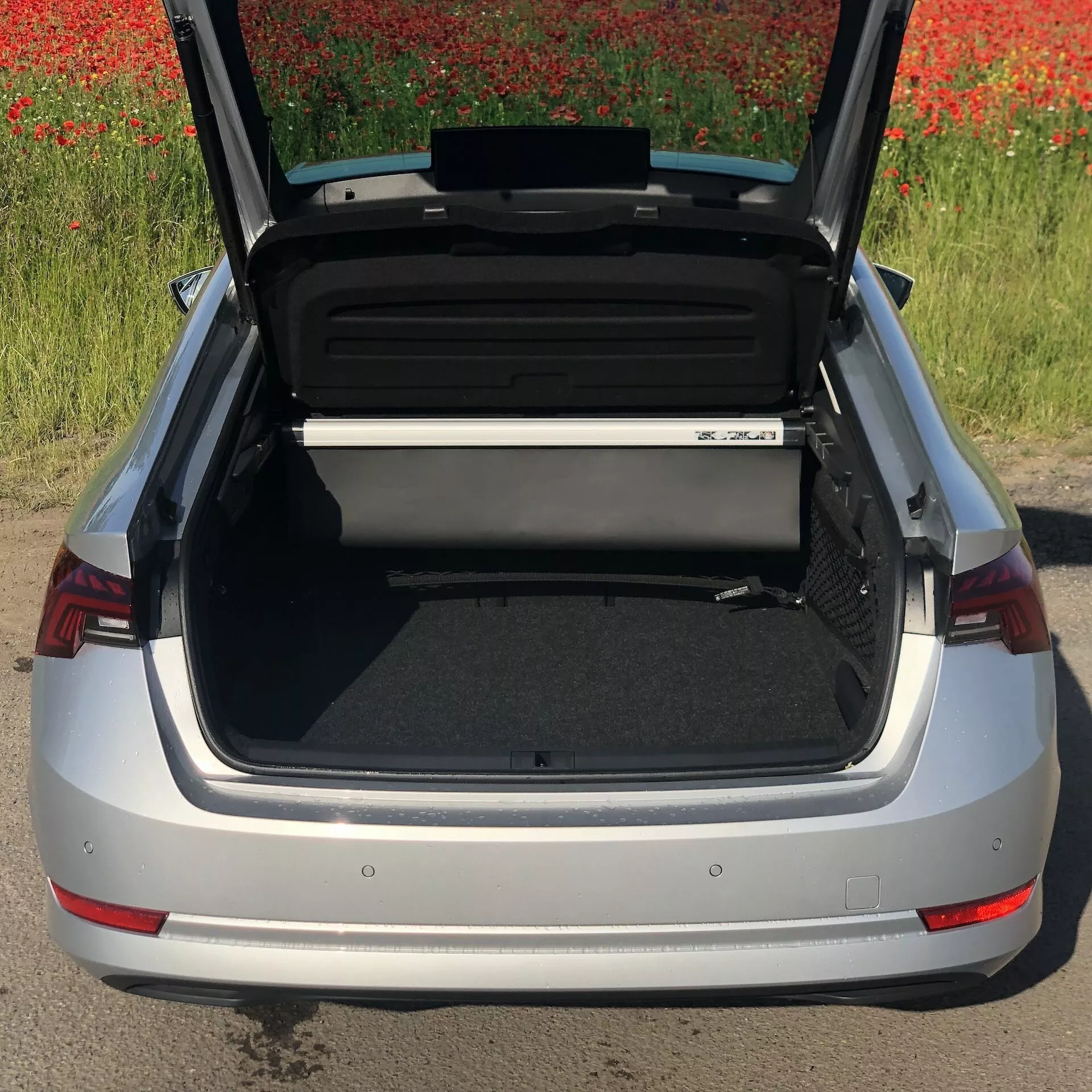 Magas szögből nézett ezüstszínű Skoda Octavia 2020 szedán nyitott csomagtartóval, amely a tágas és rendezett rakodóteret mutatja. Látható a hátsó lökhárító, a jellegzetes piros hátsó lámpák és az autó emblémájának egy része, háttérben vörös virágokkal teli mezővel.