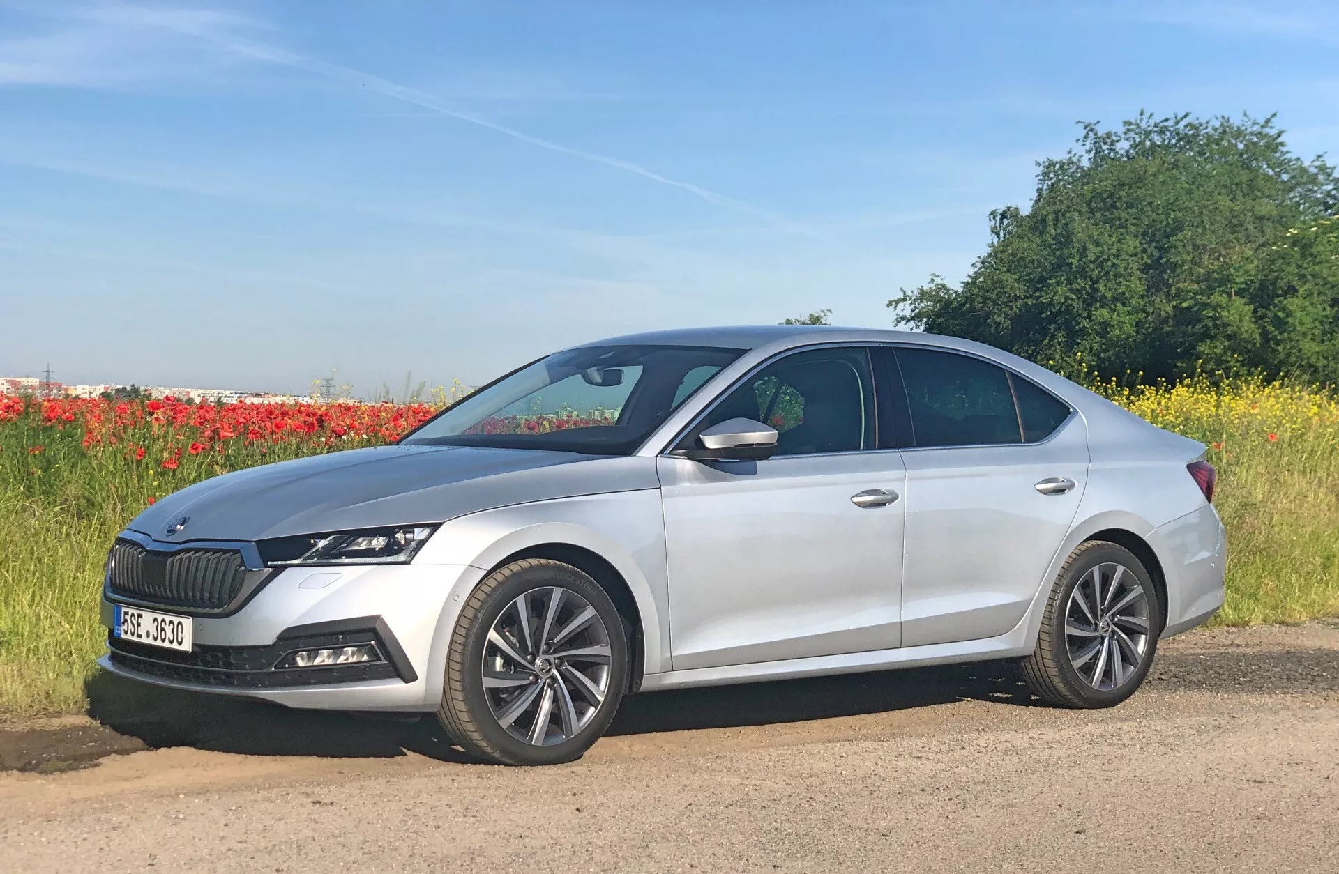 A képen egy négyajtós, ezüstszínű szedán, konkrétan a 2020-as Skoda Octavia (Octavia-Limousine) látható, egy út mellett parkolva, természeti háttérrel, amely egy mezőt és egy fasorral tartalmaz. Kissé az autó elé fordított profilról fényképezve, többrészű alumínium kerekekkel, elegáns LED fényszórókkal és kiemelkedő, függőleges lécekkel ellátott hűtőráccsal rendelkezik. A modern formatervezés magában foglalja a szobrászott karosszéria vonalakat és egy lejtős tetővonalat, integrált irányjelzőkkel rendelkező oldalsó tükrökkel. További látható jellemzők közé tartozik a hátsó antenna és az első lökhárító érzékelők, amelyek fejlett vezetői segédrendszereket jelölnek.