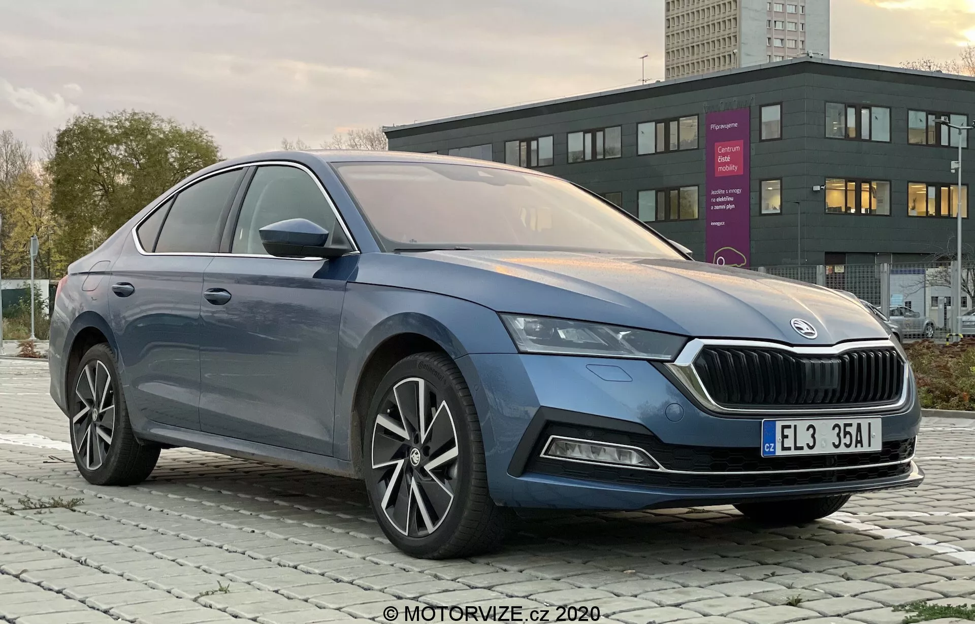 Professzionális fotó a négyajtós 2020-as Skoda Octavia Limousine-ról, amelyet elölről-balról háromnegyedes szögből készítettek, világosan mutatja a jellegzetes hűtőrácsot, elegáns LED fényszórókat, az első lökhárítót esetleges ködlámpákkal, sima karosszériavonalakat, többrudas könnyűfém keréktárcsákat, integrált irányjelzővel ellátott oldalsó visszapillantó tükröket és az ablakok prémium króm díszítését egy városi vagy külvárosi környezetben hajnalban vagy alkonyatkor.
