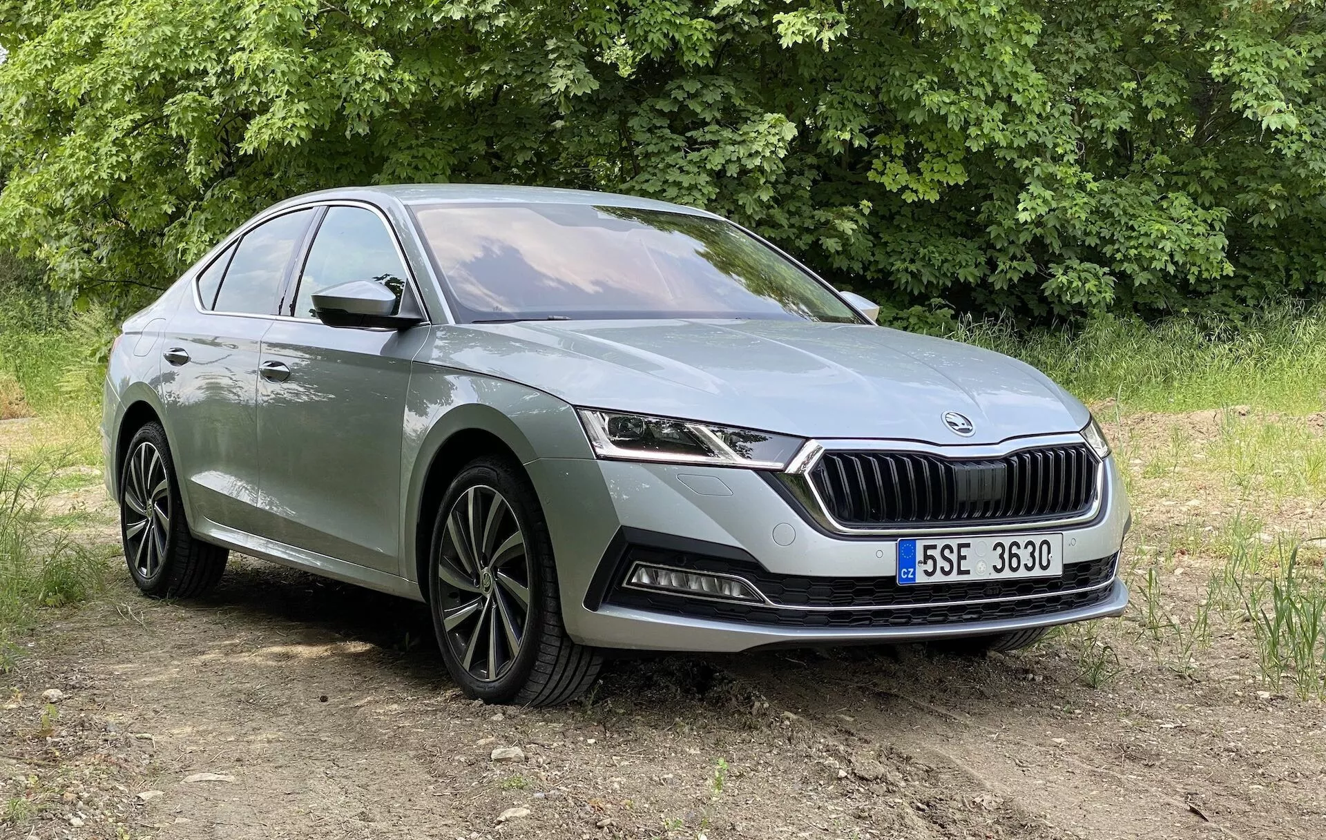 Háromnegyedes elölnézet egy modern 2020-as Skoda Octavia szedánról, kissé alacsonyabb szögből mutatva, fókuszban az első homlokrész, a függőleges rácsokkal és jellegzetes logóval ellátott hűtőrács, elegáns vízszintesen tájolt LED fényszórók, valamint az alsó lökhárító ködlámpákkal és légbeömlőkkel. Az autó egy burkolatlan területen helyezkedik el, növényzettel a háttérben, természetes nappali fényben. További látható részletek közé tartoznak a több küllős kialakítású könnyűfém felnik, a karaktervonal, amely az autó oldalán végigfut, és az ajtókilincsek. Alkalmas az autóipari dizájn és a modern járműjellemzők bemutatására kültéri környezetben.