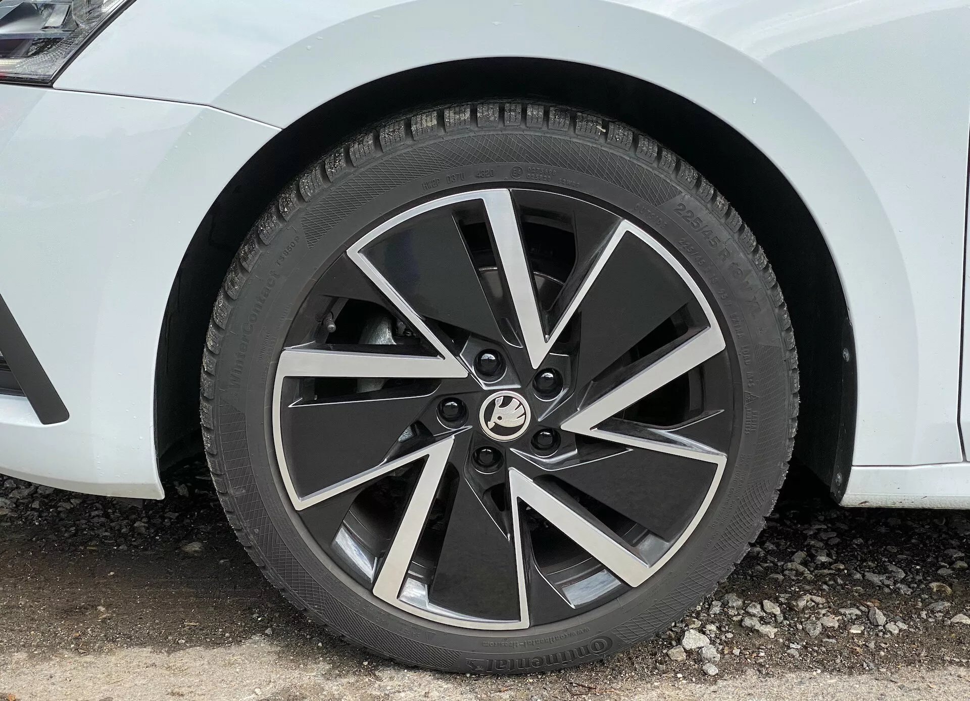 Közelkép egy 2020-as Skoda Octavia limuzin első kerekéről, modern, ötágú két tónusú felnivel és a küllőkön csiszolt felületekkel, 45 fokos szögben fényképezve, kiemelve annak dinamikus, turbinaszerű kialakítását és a négyévszakos gumiabroncs jól látható futófelületét.