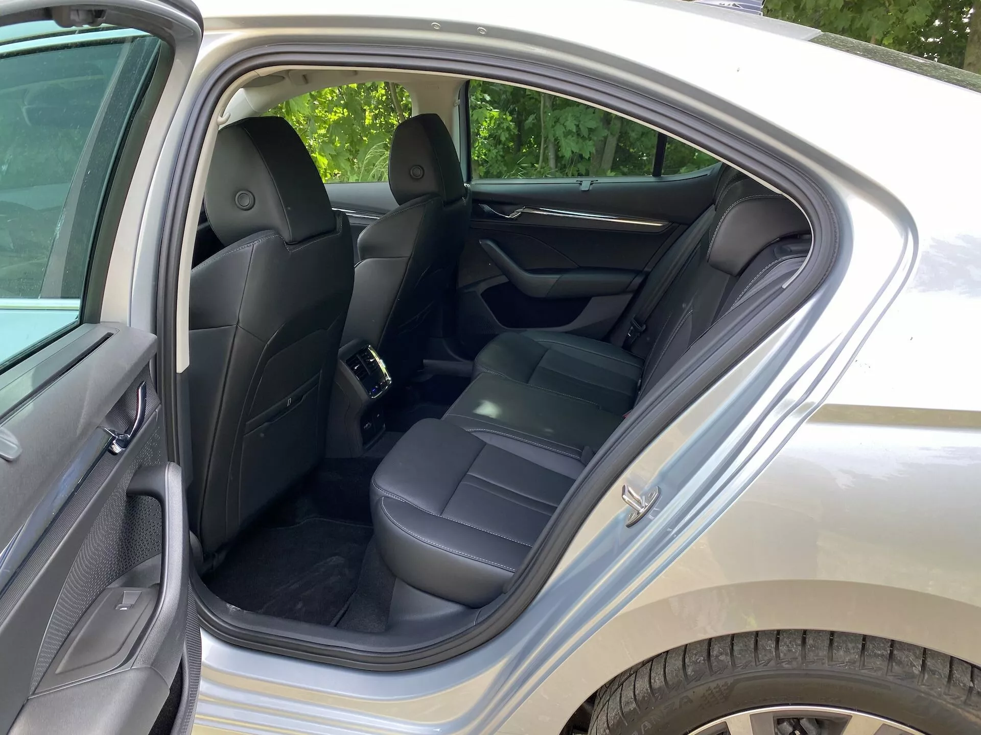 A Skoda Octavia 2020 belső terének képe, a hátsó utastérre fókuszálva. A hátsó ajtó nyitva van, megmutatva a fekete bőrrel kárpitozott üléseket, fejtámlákat, biztonsági öveket és egy felhajtott középső kartámaszt. Az ajtópanelek bőrhöz hasonló anyagból készültek a kényelem érdekében. A látható jellemzők közé tartoznak az első ülések hátulján található tároló zsebek, tiszta szőnyegek és gyermekülés rögzítési pontja. A környezeti fény azt sugallja, hogy nappal van, valószínűleg egy zöldellő külvárosi területen.