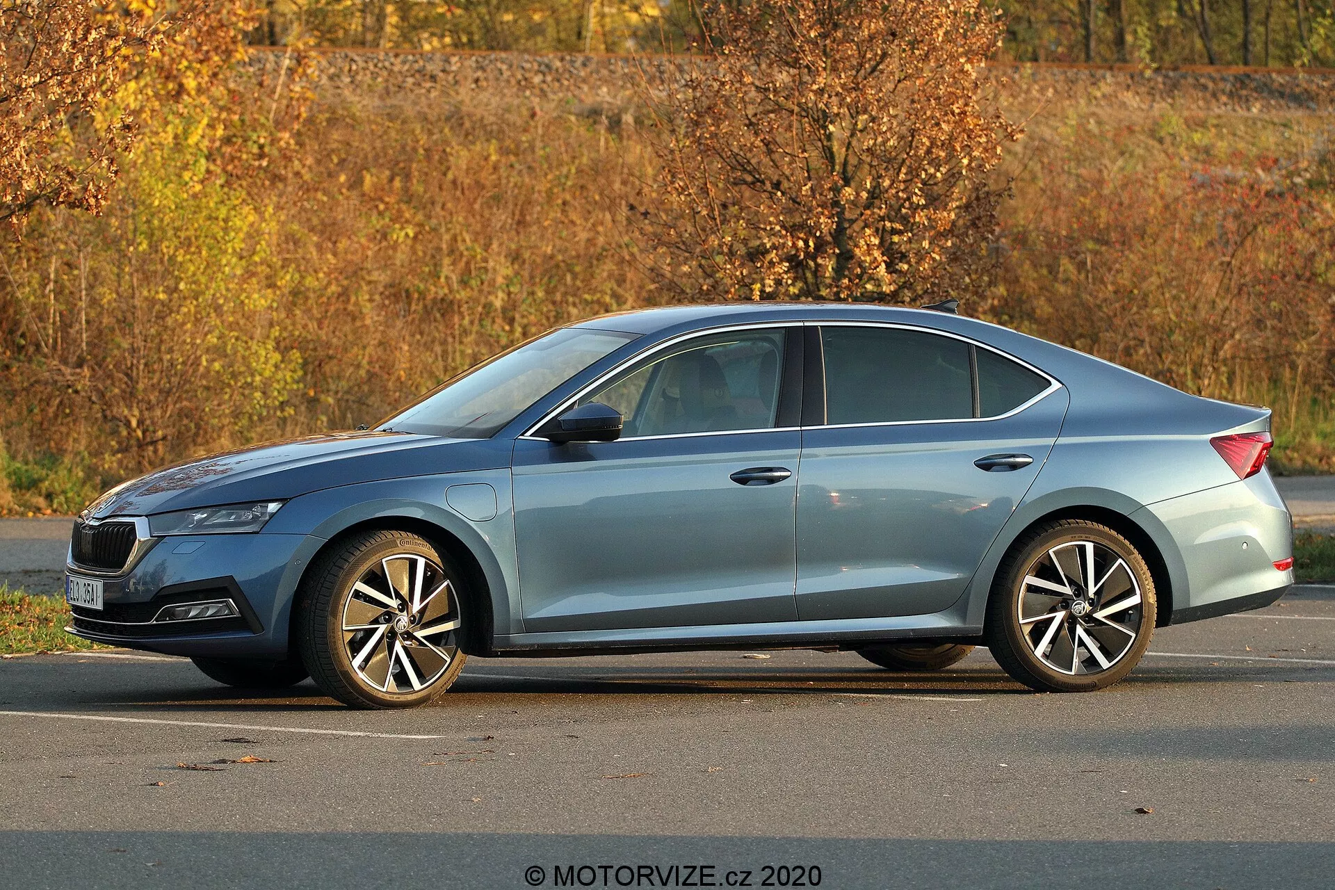 Modern Skoda Octavia 2020 sedan parkol a szabadban, háromnegyed oldalsó nézetben, teljes profil, hosszúság és magasság bemutatásával; látható részletek közé tartoznak az éles fényszórók, a lejtős tetővonal, a karaktervonalak, a krómkeretes ablakok, a többküllős alufelnik, az őszi lombhullató környezet és a természetes nappali megvilágítás.