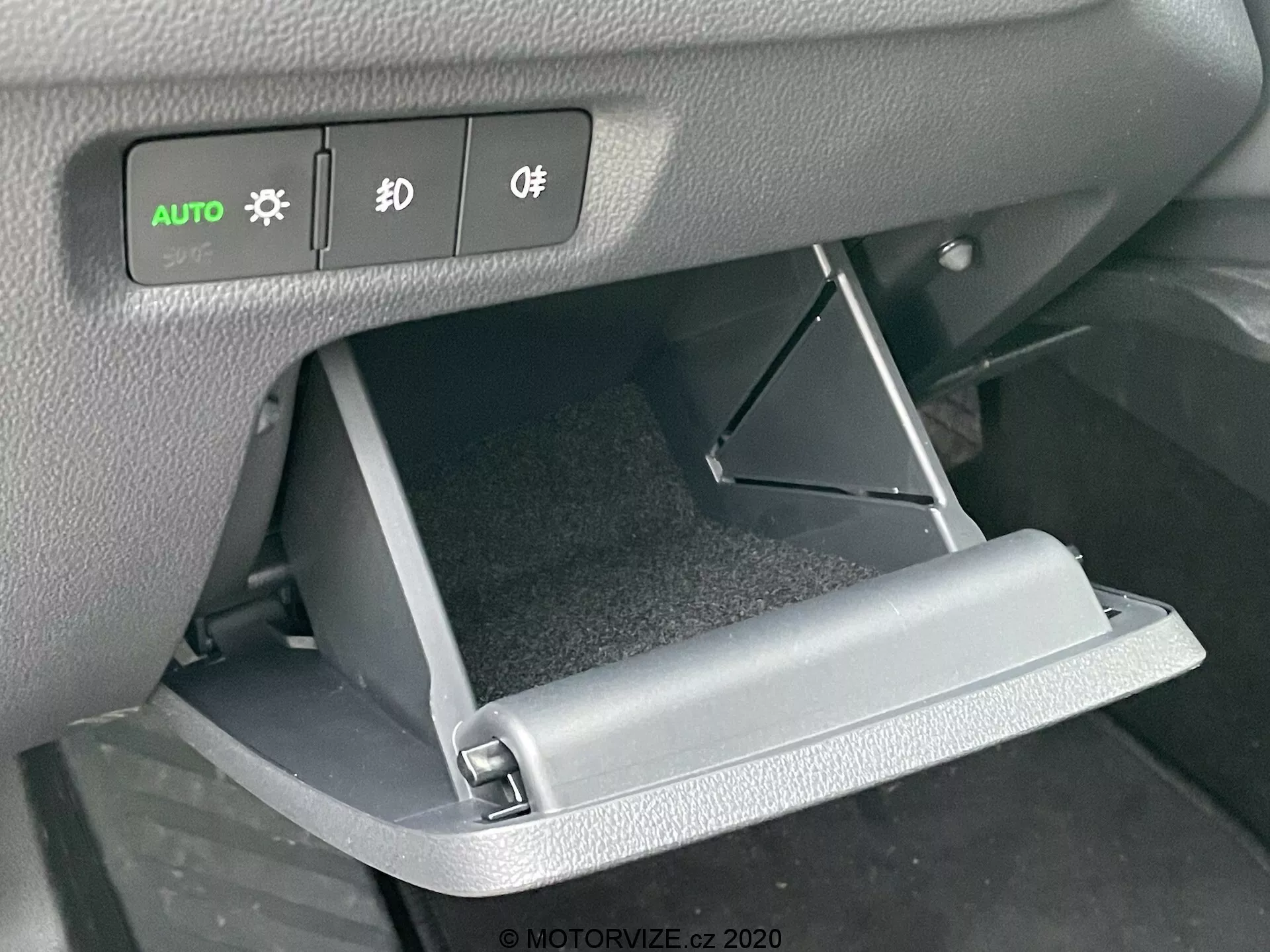 Közelkép a 2020-as Skoda Octavia belső teréről, amely a nyitott kesztyűtartóra és a műszerfal vezérlőire összpontosít. A szemből készült felvétel kiemeli a lefelé nyíló kesztyűtartó ajtaját és az alatta lévő világító fényszóró-, ködfényszóró- és világításvezérlő gombokat.