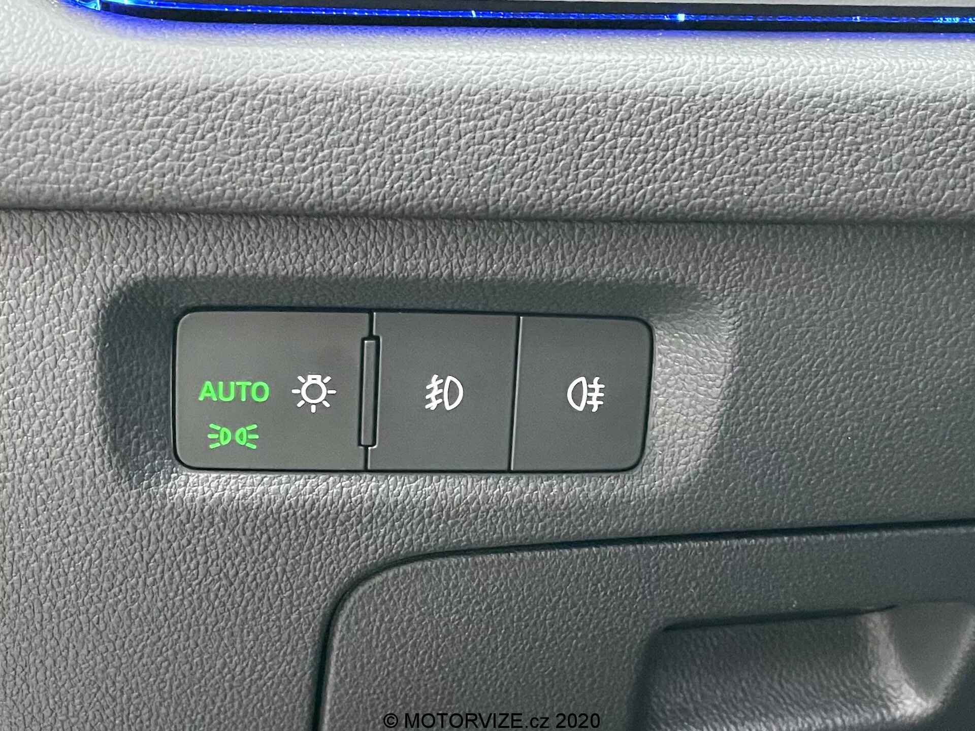 Közelről nézett Skoda Octavia 2020 vezérlőpanel, amely az automatikus fényszóró, a parkolófények és a ködlámpák kapcsolóira összpontosít. Az AUTO gomb zölden világít, jelezve az automatikus fényszóróvezérlés aktív állapotát. A texturált műanyag panel kéttónusú kialakítással rendelkezik, felül egy esetleges kék tükröződéssel, a jármű belső elrendezésében.