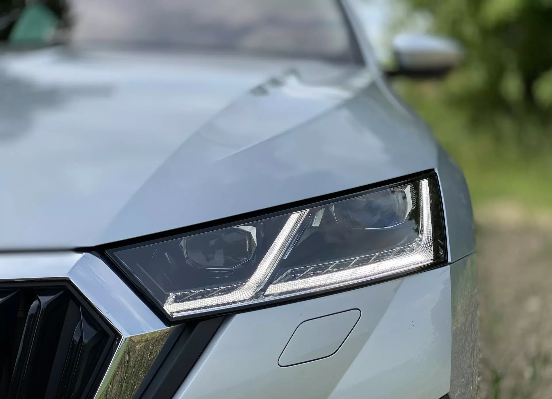 Közelkép egy modern autó bal első oldaláról, amely részletesen bemutatja a tervezési elemeket, például a szögletes LED fényszórót beépített nappali menetfénnyel, a részben látható geometriai mintázatú hűtőrácsot és a karakteres redőkkel rendelkező motorháztetőt. A magas minőségű festékbevonat és az elmosódott háttér kiemeli az autó sportos és modern esztétikáját.