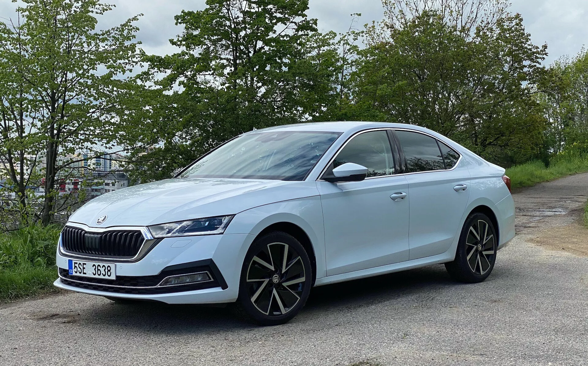 A világoskék Skoda Octavia 2020 szedán enyhén szögletes oldalnézete, amely az út szélén helyezkedik el, zöld lombozattal a háttérben. A modern dizájn jellemzi, négyajtós karosszériával, feltűnő hűtőrácsával, agresszív LED fényszórókkal, nagy, többrészes könnyűfém kerekekkel, karosszéria színű oldalsó tükrökkel, beépített irányjelzőkkel, hátsó része felé lejtő tetővonallal, sötétített üvegekkel króm díszítéssel, süllyesztett ajtónyitó kilincsekkel, alacsony profilú gumiabroncsokkal és egy jelvény, ami a modellt vagy a technológiai csomagot jelzi.