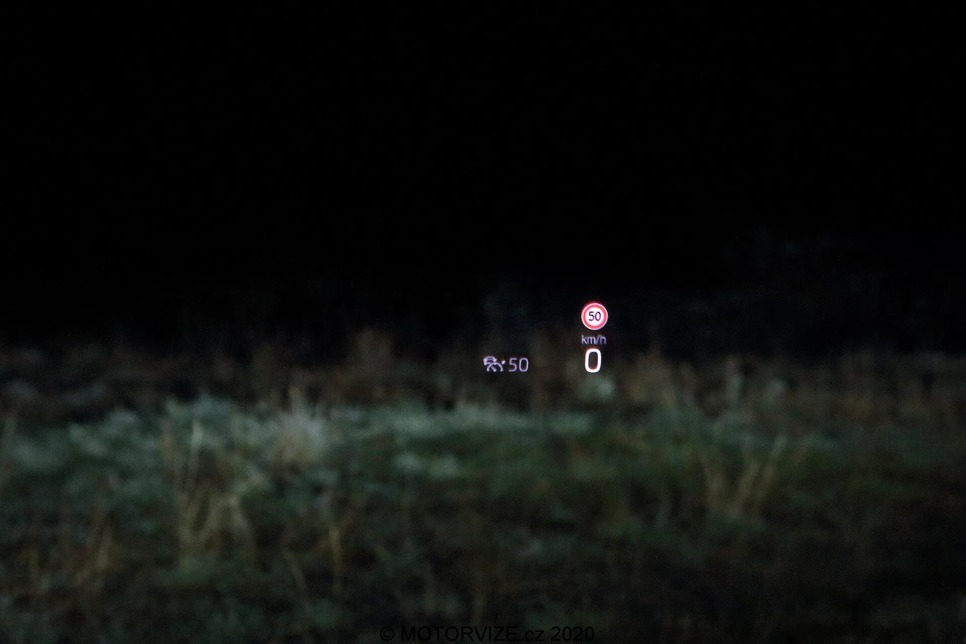 Kép, amely bemutatja a Škoda Octavia 2020 limuzin head-up display (HUD) technológiáját. A kijelző a szélvédőre van vetítve, és 0 km/h sebességet mutató álló sebességmérőt, valamint egy tiltó jelet jelző ikont mutat, amely egy nem engedélyezett funkcióra utal, elmosódott növények hátterében.