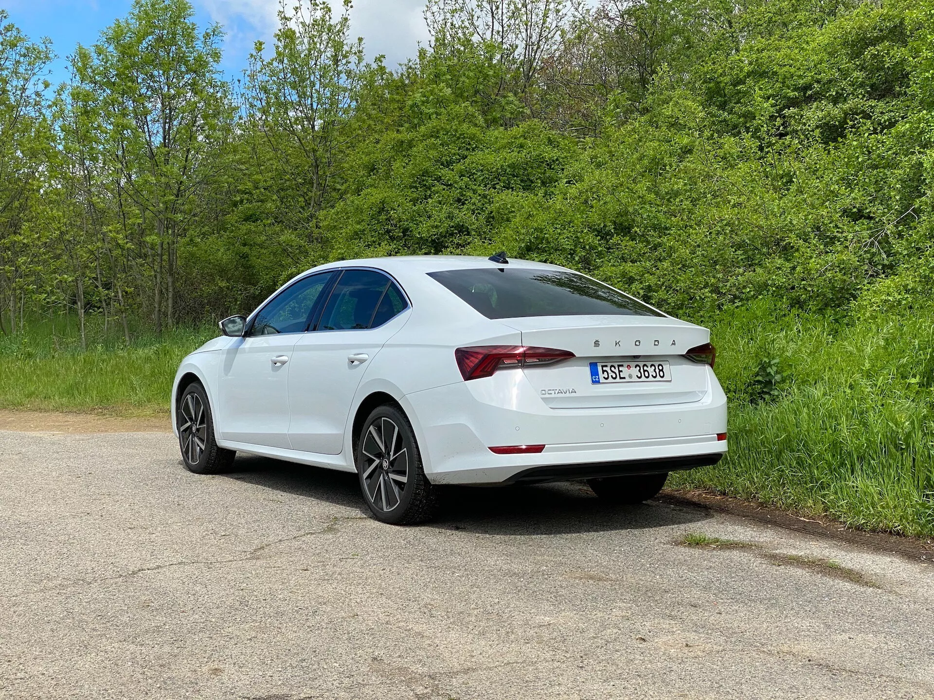 2020-as Skoda Octavia fehér szedán háromnegyed hátsó nézetben, kavicsos úton parkolva, növényzet övezte területen, részletes nézetben megjelenítve a jellegzetes hátsó lámpákat, a hátsó fedélszárnyat, az alufelniket és a látható napfénytetőt, kiemelve modern kialakítását és technológiai jellemzőit, beleértve a lökhárítón elhelyezkedő parkoló szenzort.